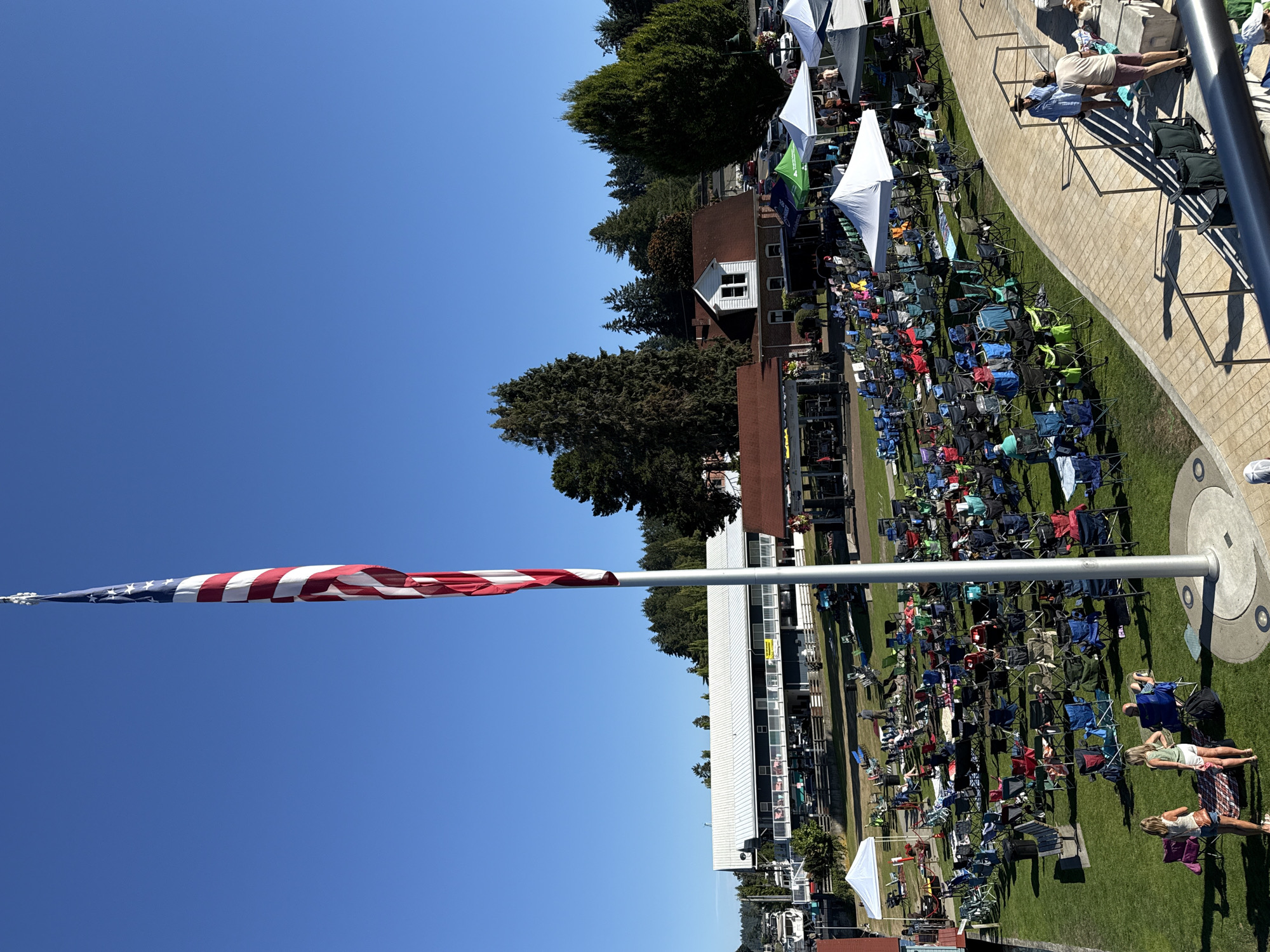 In Gig Harbor, Washington, a large American flag on a tall flagpole stands prominently against a clear blue sky. The scene below depicts an outdoor gathering, likely an event or concert, with numerous folding chairs set up on a grassy area. Several white and green tents are also visible. People are seated in the chairs, and others are standing and interacting. A long, white building with a silver roof is situated in the background, with boats docked nearby. The overall atmosphere suggests a sunny day and a community event.