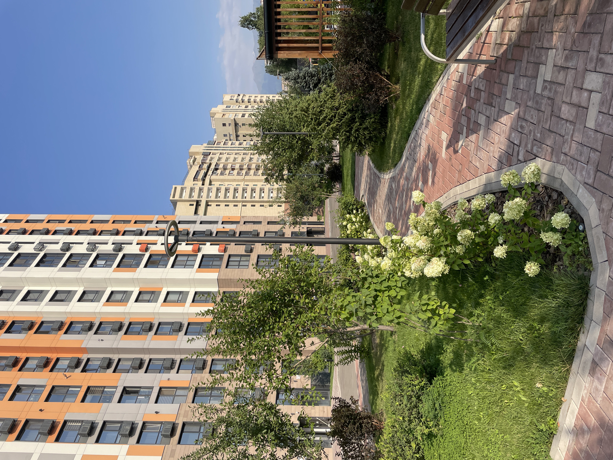 A low-angle outdoor shot captures a paved walkway in a city park area, bordered by manicured green lawns and various trees and shrubs. The walkway, made of red and grey bricks laid in a herringbone pattern, curves to the right, leading the eye past flowering bushes with white blooms. To the left, a tall residential building with a facade of orange and grey panels rises against a clear blue sky. The building has numerous windows and air conditioning units. In the background, other multi-story apartment buildings with light-colored facades are visible, and distant mountains can be seen on the horizon. A wooden gazebo-like structure with slatted walls is partially visible on the right, and a dark wooden park bench with metal legs sits beside the walkway, casting a shadow. The lighting suggests it is a sunny day, possibly late morning or early afternoon, with long shadows. The scene is devoid of people.