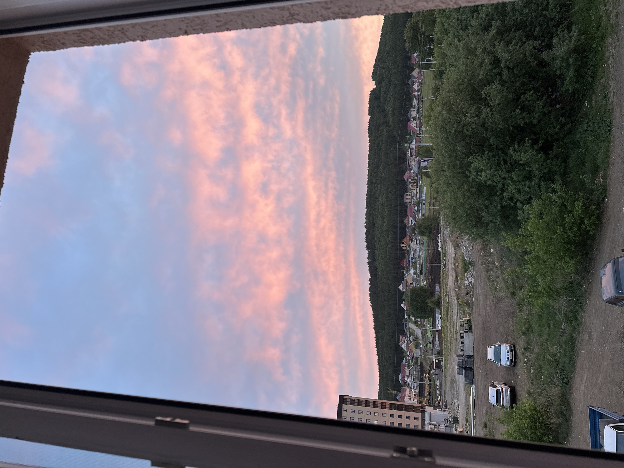 View from a window overlooking a town. The sky is filled with pink-tinged clouds. In the distance, there's a tree-covered hill and a settlement with houses. Below, there's a building, vehicles, and a dirt road.
Location: Unknown, Unknown.