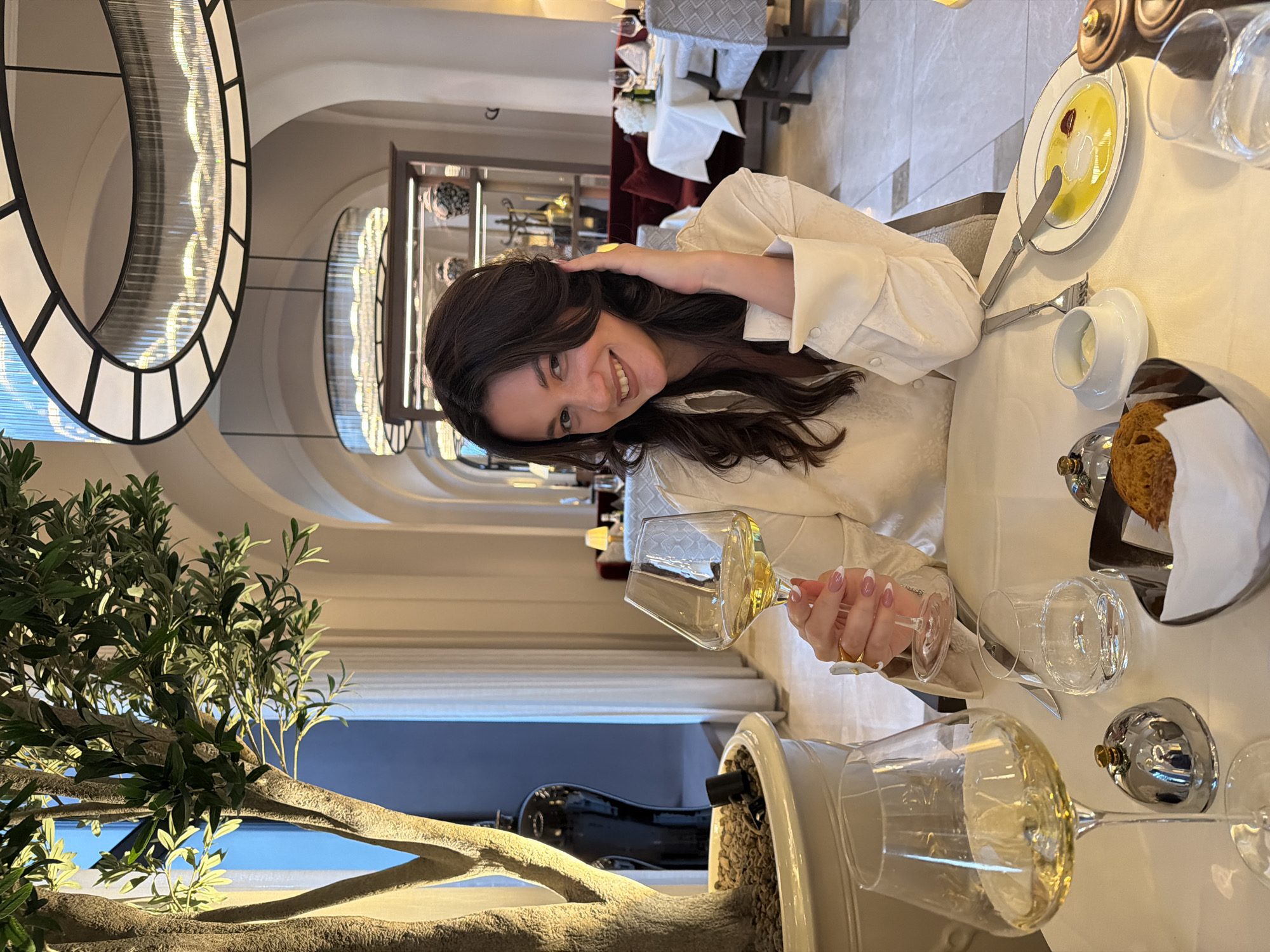 The image depicts a woman smiling and holding a glass of white wine in what appears to be a restaurant in Belgrade, Serbia. She is seated at a table set with plates, bread, glasses, and cutlery. Behind her is a hallway with arched openings and a decorative shelving unit. A potted tree is placed in the foreground.