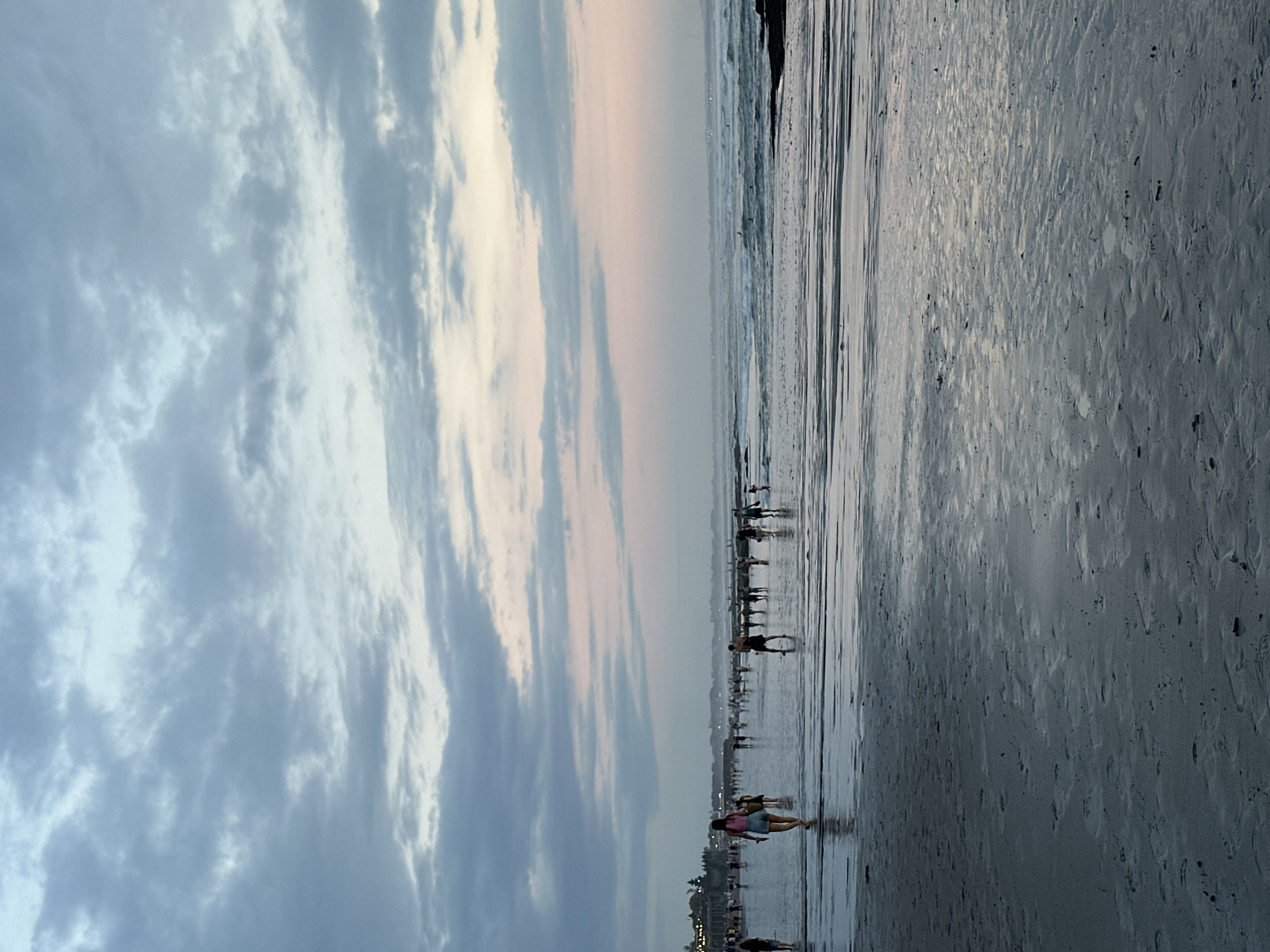 The image depicts a beach scene in North Kuta, Indonesia, at dusk. A wide sandy beach occupies the right half of the frame. People are walking along the shoreline, their silhouettes visible against the wet sand reflecting the sky. The ocean is to the left of the beach, with gentle waves coming to shore. The sky dominates the left half, filled with layered clouds displaying varying shades of gray, white, and a hint of pink from the setting sun. A faint glow is visible on the horizon, suggesting a sunset. A cityscape is dimly seen in the far distance.
