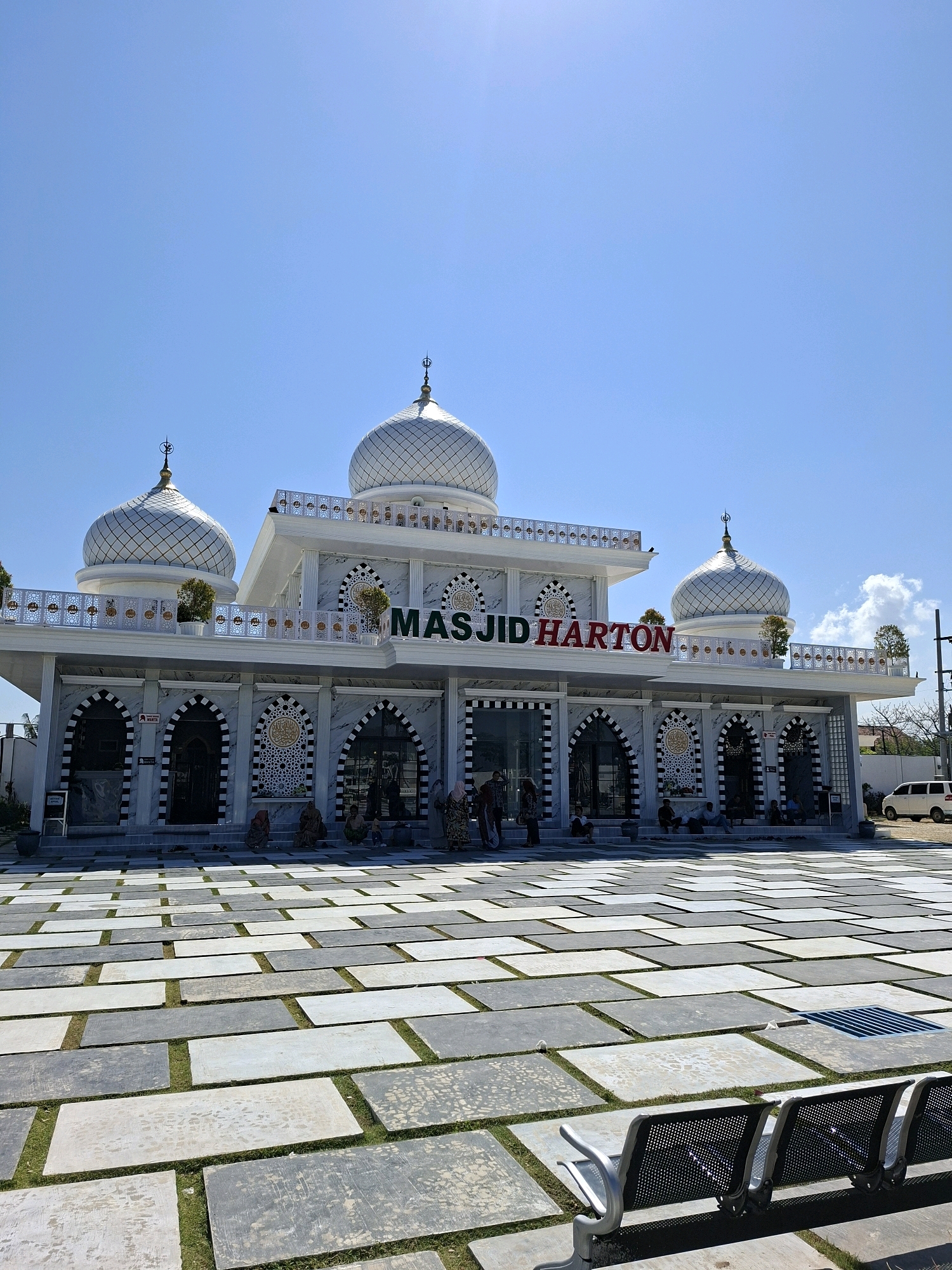 The image shows the Masjid Harton, a white and marble mosque located in Camplong, Indonesia. The mosque features several domes, ornate decorative panels, and the name "MASJID HARTON" in red lettering above the entrance. The foreground is a paved courtyard with rectangular grey and white tiles interspersed with grass. Several people are seated on the steps of the mosque and walking around the courtyard. In the background, a white van is parked to the right of the mosque. The sky is clear and blue.