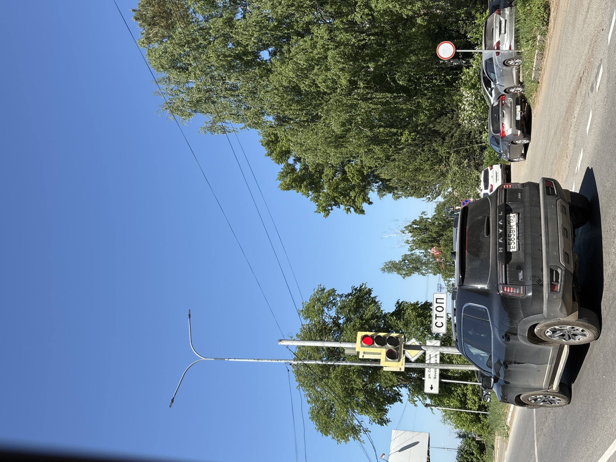 The image shows a street scene in Chaikovsky, Russia, with a black Haval Jolion SUV stopped at a red traffic light. A "STOP" sign in Russian and a street sign for Sovetskaya Street are visible near the traffic light. Several parked cars line the right side of the road. Overhead power lines are present, and trees are in the background against a clear blue sky.