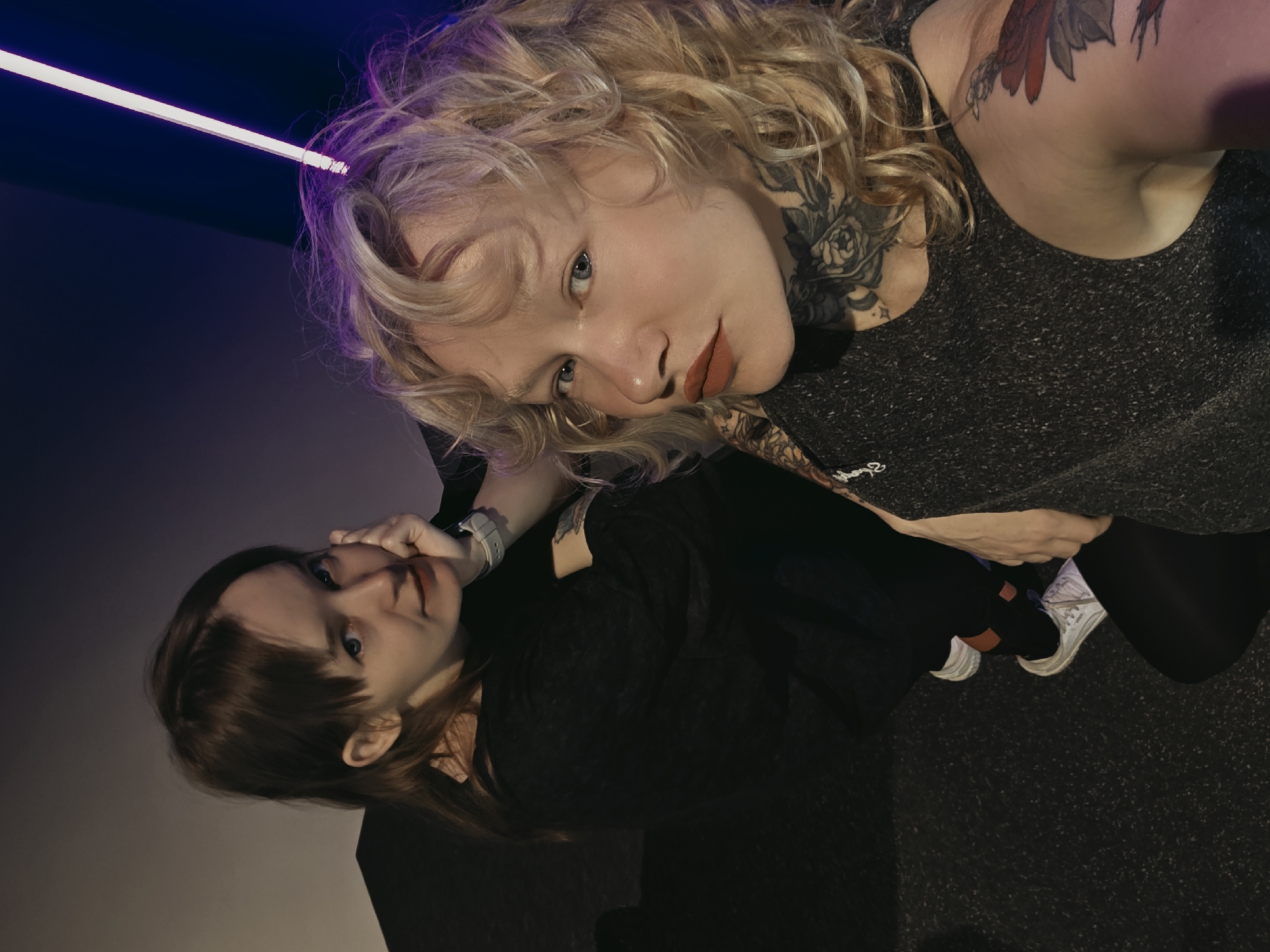 Two women are positioned in what appears to be an indoor, dimly lit environment, possibly a club or event space. The woman on the left has brown hair with bangs and is wearing a black long-sleeved shirt and black leggings, visible white sneakers with orange socks. She is seated, with her left hand resting on her cheek and her right arm bent, her hand near her face. The woman on the right has blonde, curly hair and is wearing a dark grey sleeveless top. She has extensive tattoos on her neck, chest, and arm. She is also seated, leaning forward. A neon light fixture, emitting a purple glow, is visible in the background, casting a colored light on the scene. The floor is dark and textured, resembling carpet. The background wall is a gradient from light grey to dark purple.