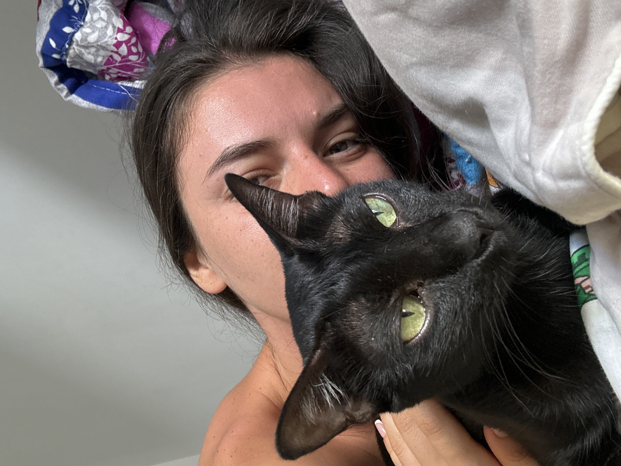A young woman is pictured in a close-up shot with a black cat. The woman has dark hair and fair skin with freckles. She is smiling slightly and looking at the camera. The black cat has green eyes and is being held by the woman's hand, which is visible in the lower right corner of the frame. The background includes a white sheet and a colorful patterned pillow. The location cannot be determined from the image alone.