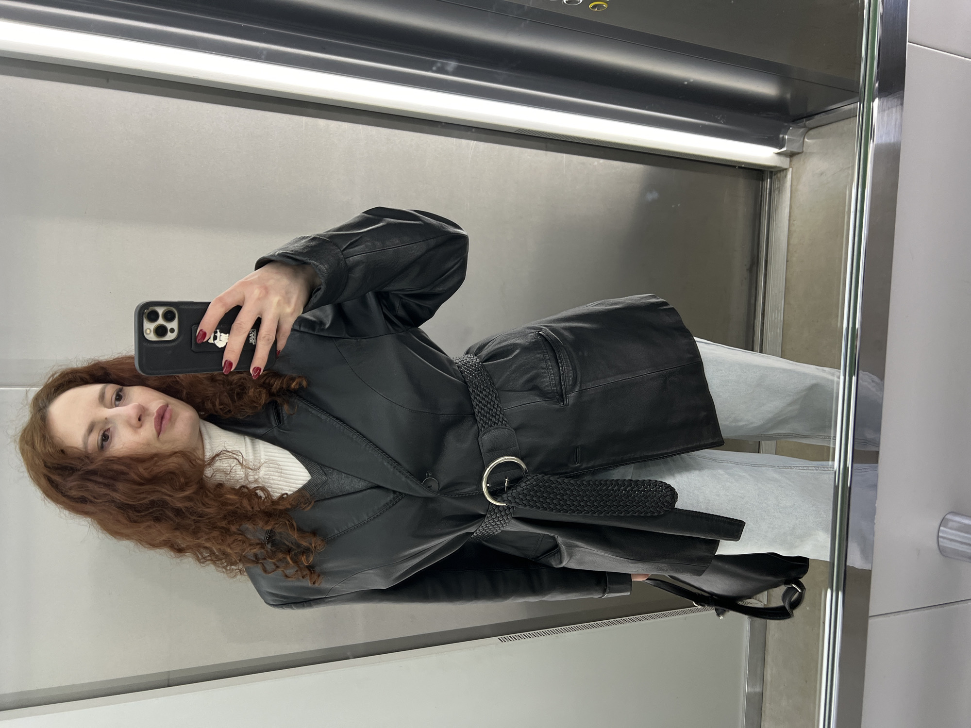 A red-haired woman is taking a selfie in a mirror inside a metallic elevator. She is wearing a black leather jacket with a braided belt, a white turtleneck sweater, and light blue jeans. The mirror's reflection shows the elevator's interior: metallic walls, a bright overhead light, and a door on the right side of the frame. The woman's phone is a black smartphone with multiple camera lenses, and her fingernails are painted red. The background consists of metallic panels that appear to be part of the elevator's interior. There is no indication of Saint Petersburg.
