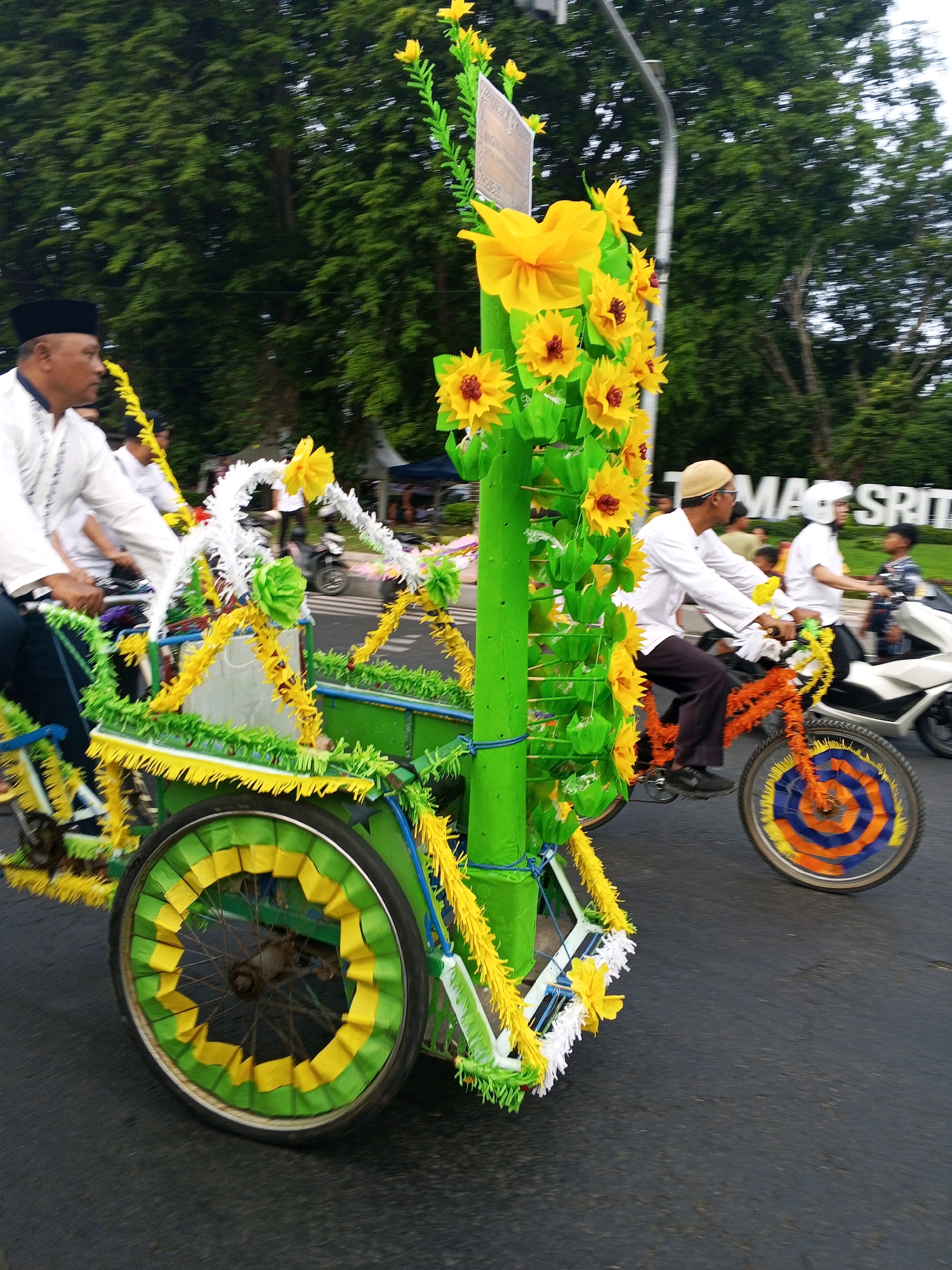 This is a medium-shot, eye-level, outdoor, daytime photograph of a parade or procession in Banyuwangi, Indonesia. The foreground is dominated by a brightly decorated pedicab, with its front wheel and side visible. The pedicab is adorned with green and yellow paper flowers and streamers. A person wearing a black peci hat and a white shirt is partially visible on the left, seemingly part of the procession. In the mid-ground, a man wearing a white shirt and a tan skullcap rides a bicycle with an orange frame, which is also decorated with colorful paper elements, including on its wheel. To the right of the cyclist, another person on a motorbike is visible. In the background, lush green trees line the street, and a large sign displaying "MAG SRIT" is visible. The overall impression is one of a festive, community event.