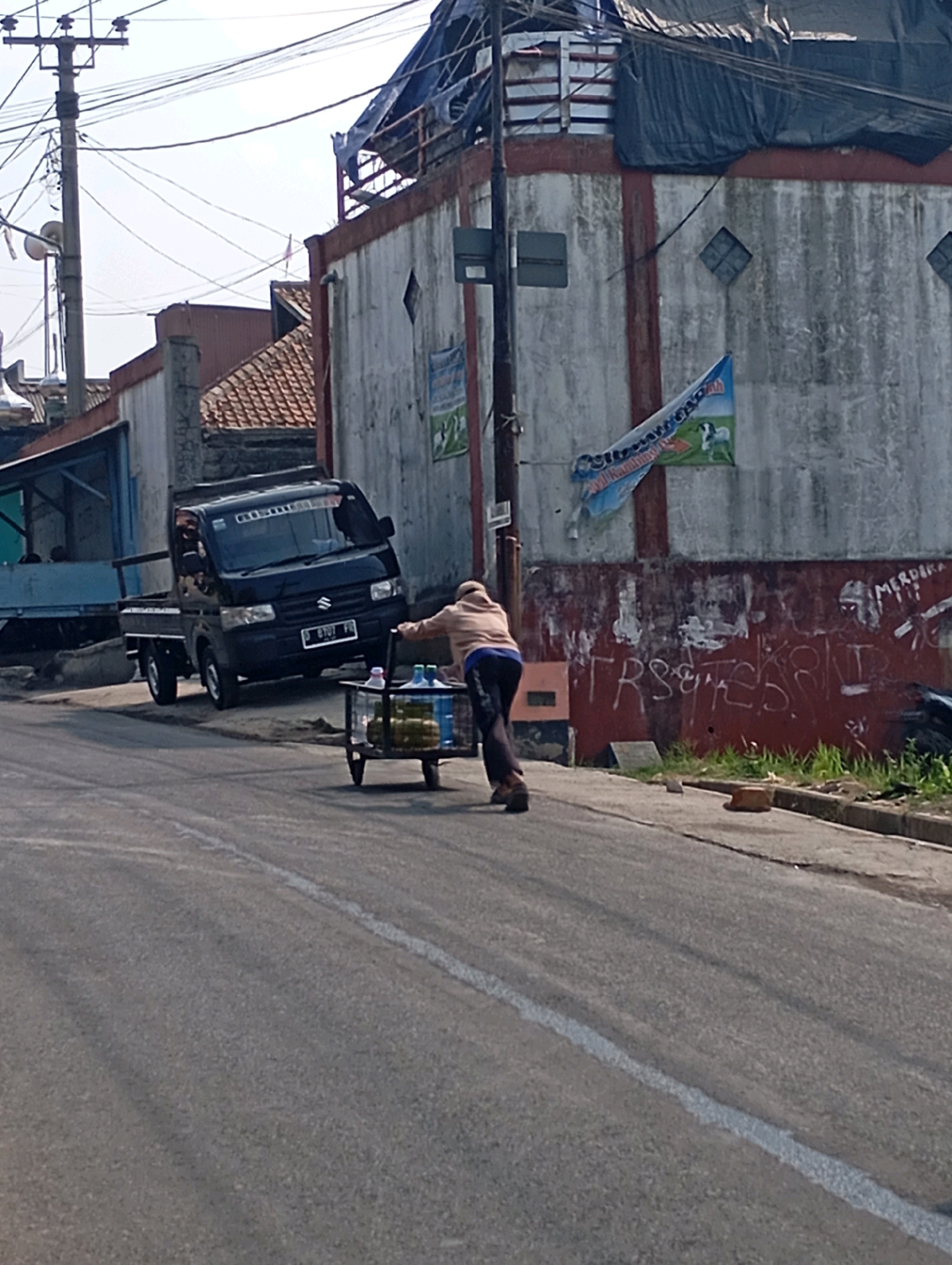 A person is pushing a cart filled with bottled drinks down a paved road in Cimahi, Indonesia. The person is wearing a light brown jacket and dark pants, and the cart has a metal frame with two wheels. In the background, a black Suzuki pickup truck is parked on the side of the road. A dilapidated building with graffiti and a tattered blue tarp covering part of its roof is visible behind the truck. Electrical wires and poles crisscross the sky, typical of urban landscapes in Indonesia. The road appears to be a local street with some debris and grass growing along the roadside. The scene suggests a vendor moving their goods through the city.
