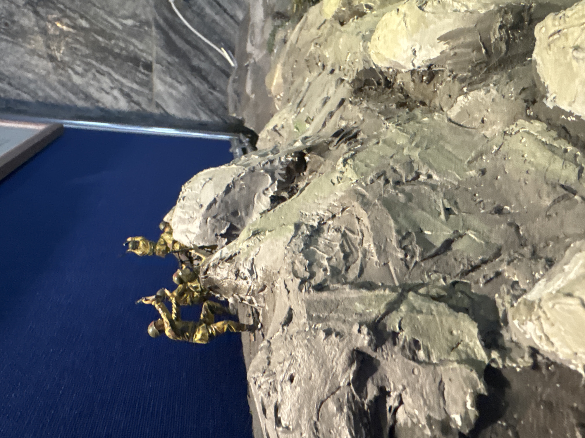 A close-up photograph depicts a diorama of soldiers climbing a rocky terrain. The figures are small, and their uniforms are shaded in green and brown tones. The rocky landscape is made of textured gray and light beige material, giving it a sculpted appearance. A deep blue background is visible behind the diorama on the left side of the image. It is located in the Siege of Leningrad Museum in Saint Petersburg, Russia.