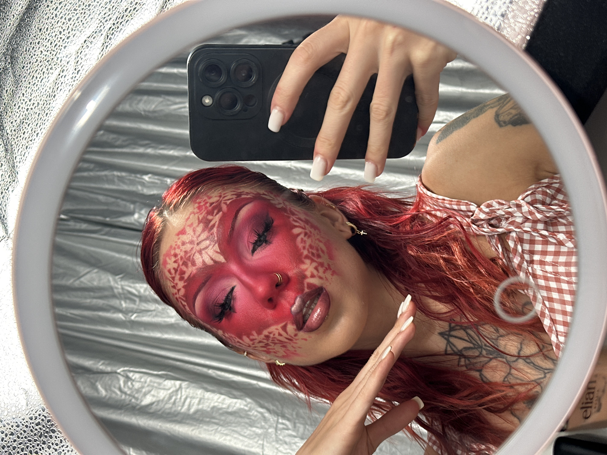 This is a first-person perspective media file, likely taken in North Kuta, Indonesia. The subject is a person, reflected in a ring light mirror, who is taking a selfie with a smartphone.
The person has vibrant red hair and dramatic makeup on their face, featuring a red base with intricate white lace-like patterns extending from the forehead and cheeks. They are wearing dark eyeshadow, false eyelashes, and metallic lipstick. Gold jewelry, including a nose ring and hoop earrings, is visible.
The person is wearing a red and white checkered top. Tattoos are visible on their shoulder and upper chest, with one tattoo on the chest appearing to be a geometric floral design. Their fingernails are painted white.
The background is a crinkled silver fabric, suggesting a studio or indoor setting. The lighting appears bright, likely from the ring light itself, creating a soft glow. The overall mood is artistic and self-expressive.
No specific time of day or weather conditions can be determined from the image. No visible text is present in the frame.