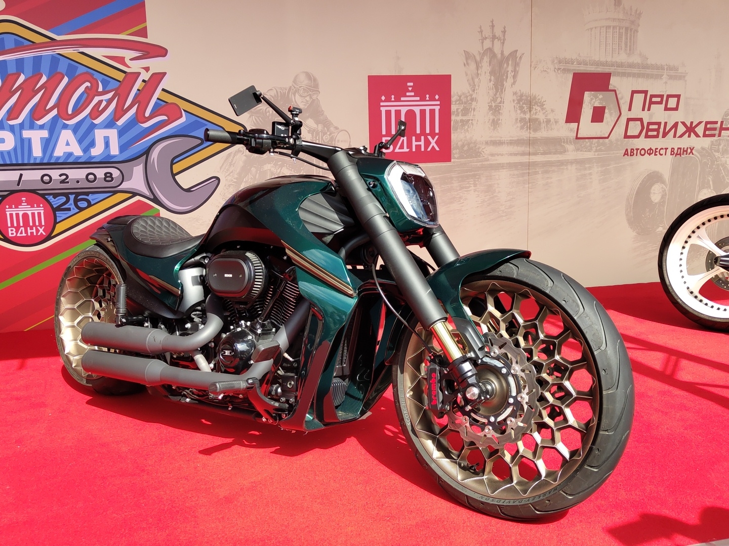 The image displays a custom-built, dark green motorcycle parked on a red carpet. The motorcycle has a distinctive wheel design with a hexagonal pattern. In the background, there's a large banner with text and graphics, including what appears to be a wrench and buildings. The banner also features Russian text, with "ВДНХ" (VDNKh) clearly visible, indicating the location as VDNKh (Exhibition of Achievements of National Economy) in Moscow, Russia. The overall scene suggests an exhibition or show.