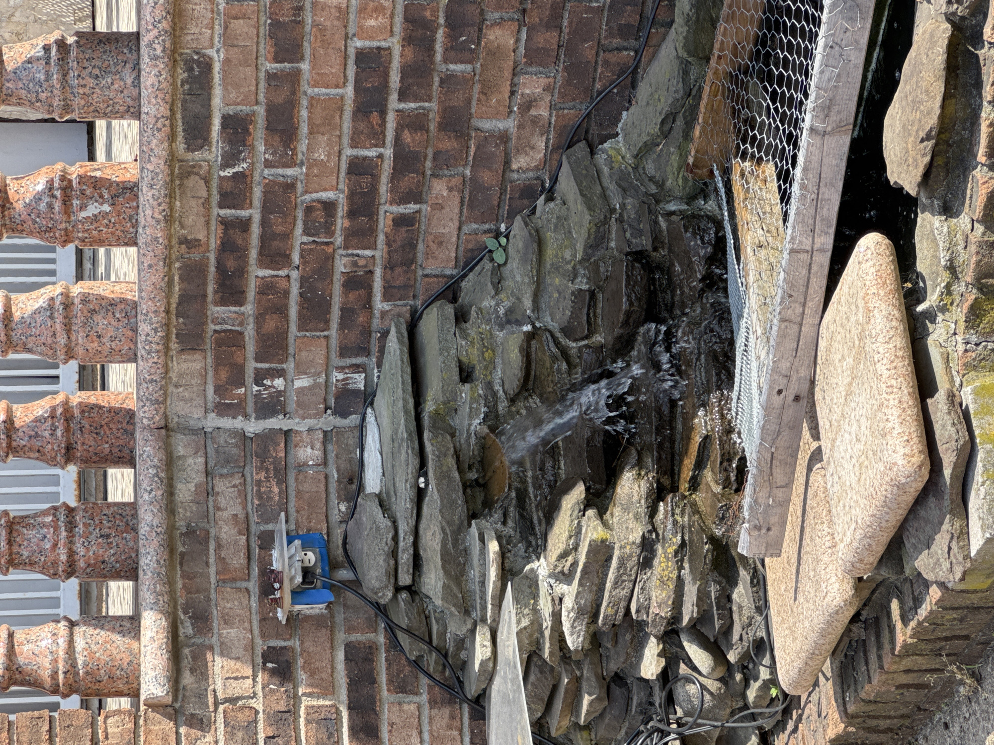 The image shows a close-up, eye-level view of an outdoor water feature against a brick wall. The water feature is constructed from stacked rocks, with water cascading down some of the rocks. A black electrical cord snakes along the wall and rocks, leading to a blue electrical outlet box mounted on the brick. Three decorative granite balusters are visible at the top of the frame, set against a white railing. In the foreground, large flat stones rest on a brick base, with a wooden frame covered in wire mesh partially submerged in dark water. There are no people or animals visible in the image. The scene appears to be in an outdoor setting, possibly a backyard or garden. The location in the city of New York, United States of America is not discernible from the visual information present in the image.