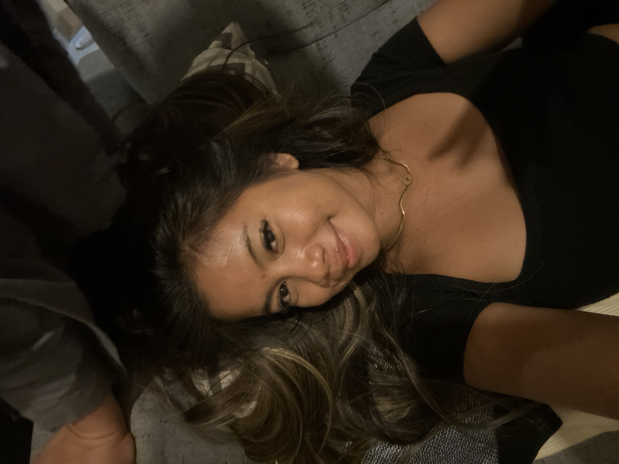 A woman with dark hair and brown highlights is lying on a gray sofa. She is wearing a black top and a gold necklace. The background is blurred, but appears to be an indoor setting. There is no information in the image to determine its location in Central Jakarta, Indonesia.