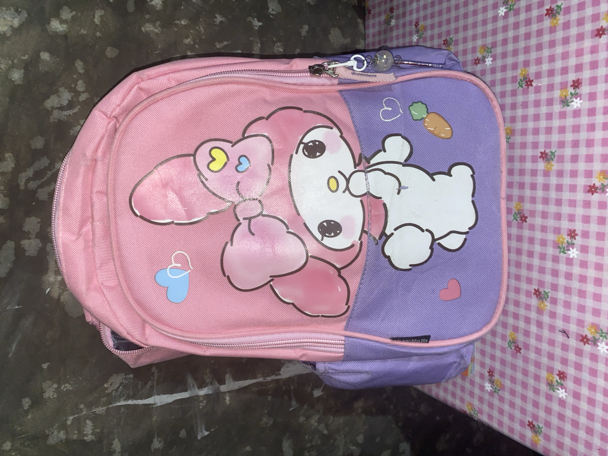 A pink and purple backpack featuring the character My Melody is displayed against a textured, dark background. The backpack has a large graphic of My Melody with a pink bow and is adorned with small hearts and a carrot. A zipper pull with a clear bead is attached to the side zipper. The backpack rests on a pink and white checkered surface with scattered floral patterns. The overall scene suggests a child's item or accessory. The background's texture hints at an indoor setting, possibly a wall, and the patterned surface implies a table or shelf. The image does not provide information about its location in Bontoharu, Indonesia.