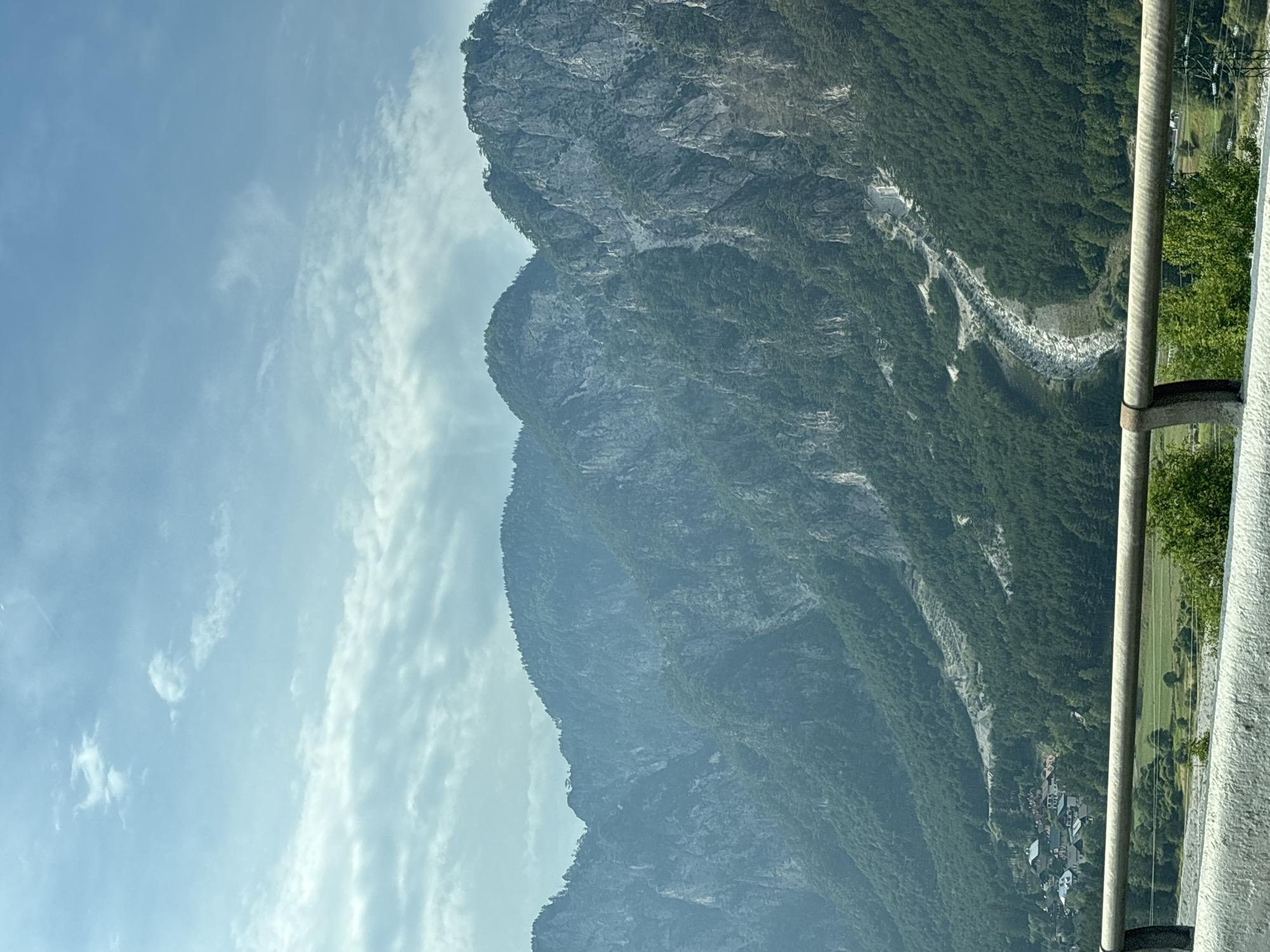 This is a distant, high-angle shot of a mountainous region, likely in Italy given the mention of Malborghetto-Valbruna. The scene is dominated by lush green forests that cover the slopes of imposing, rugged mountains. The mountains themselves appear steep and rocky, with lighter grey patches suggesting exposed rock faces or scree slopes. A visible dry riverbed or trail snakes its way down one of the mountain slopes, cutting through the dense greenery. In the lower left portion of the image, a small village with red-tiled roofs is nestled among the trees, indicating human habitation in this natural landscape. The foreground is partially obstructed by a concrete barrier and a metal railing, suggesting the photo was taken from an elevated viewpoint, possibly a road or a bridge. The sky above is a pale blue with scattered white clouds, some of which are wispy and streaky, possibly indicating contrails. The overall impression is one of a serene and vast natural landscape with a small human settlement integrated into it.