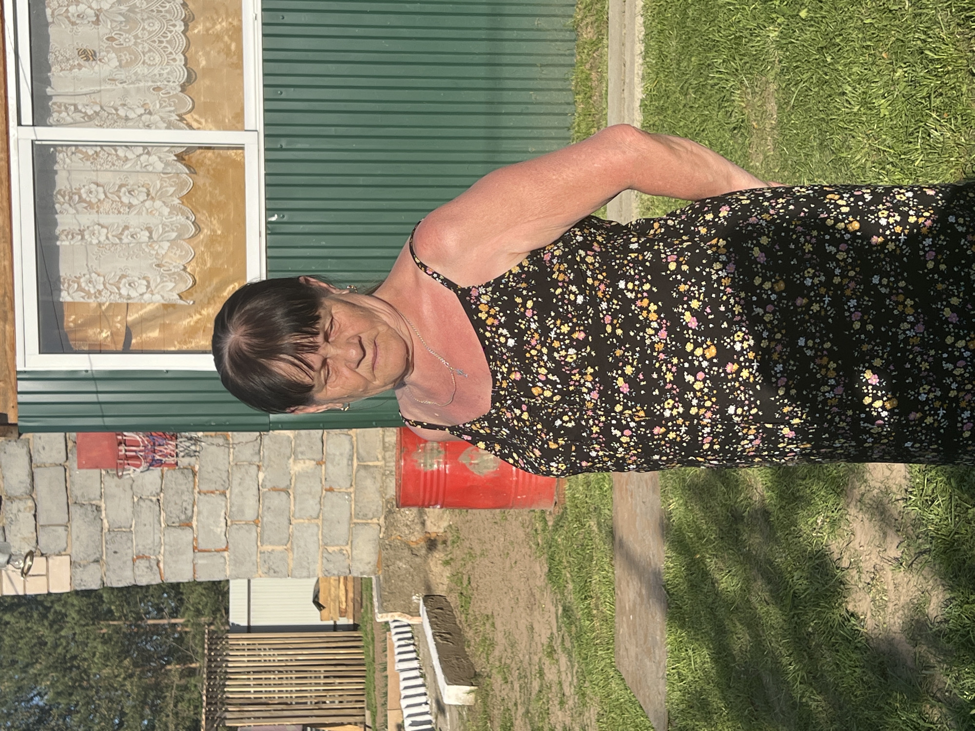 An elderly woman stands in the yard of a property. She is wearing a dark, floral-patterned dress with spaghetti straps and a silver necklace. The yard is covered in grass. Behind her is a green-sided building with a window adorned with lace curtains. Adjacent is a brick wall with a basketball hoop attached. A red barrel stands nearby. To the side is a stack of lumber. The setting appears to be in a rural or suburban area. No evidence confirms it to be in the specific city of Chaikovsky, Russia. The description is based on the objects and environment depicted in the image.