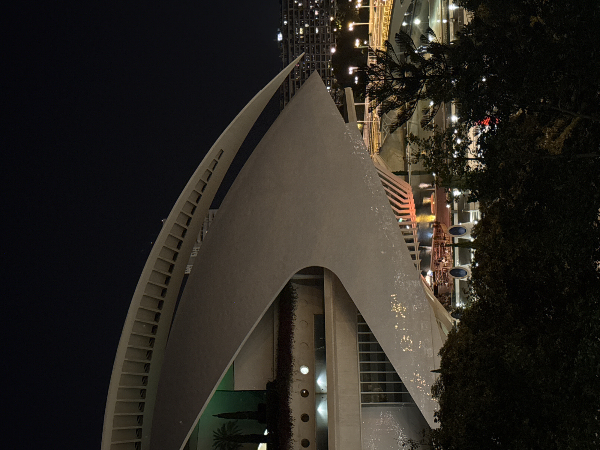 This is a nighttime photograph of the City of Arts and Sciences in Valencia, Spain, specifically focusing on the Hemisfèric building. The structure's distinctive eye-shaped design is prominent, with its white, curved facade illuminated. The dark sky serves as a backdrop, contrasted by the artificial lights emanating from the surrounding urban landscape. In the foreground, dark foliage partially obscures the view, suggesting the photograph was taken from a vantage point within a park or garden area. Lights from buildings and street lamps create a bokeh effect in the middle ground, hinting at the presence of activity within the complex, such as outdoor dining or events. The overall impression is one of modern architecture set against the vibrant energy of a city at night.