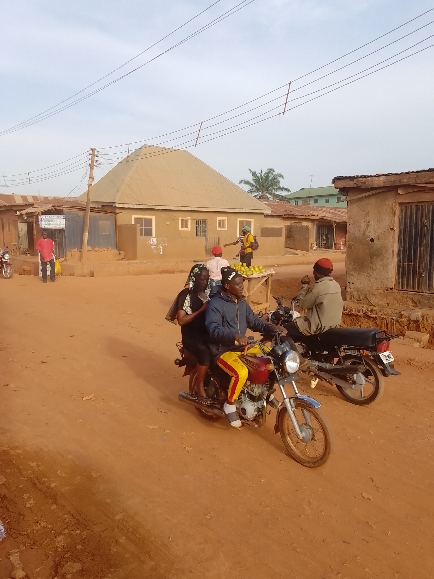 This image captures a vibrant street scene in Yelwa, Nigeria, under a clear, bright sky, suggesting late morning or early afternoon. The ground is unpaved, reddish-brown dirt, indicating a rural or semi-urban setting.
In the foreground, a person wearing a dark jacket and yellow and red striped pants (with "LAKERS" visible) rides a red and yellow motorcycle. A passenger in a dark top and patterned head covering sits behind them. To their right, another individual in a light-colored jacket and red cap is seated on a dark motorcycle, facing away from the camera. The dark motorcycle's license plate displays "PNK" and "2K".
Further into the background, several people are visible, including a person in a yellow shirt standing next to a wooden stall laden with bright green fruits, likely for sale. Another person in a red shirt holds a yellow bag nearby. The surrounding buildings are modest, with various styles including a prominent structure with a light-colored, conical or dome-shaped roof. Utility poles and overhead power lines crisscross the upper part of the frame, and a palm tree is visible in the distance. On a blue stall to the left, partially visible text reads "DAVE SACRAMENTAL" and a phone number starting with "0706...".
The scene depicts everyday activities, with people riding motorcycles and engaging in street vending, creating a typical, bustling atmosphere of daily life.
