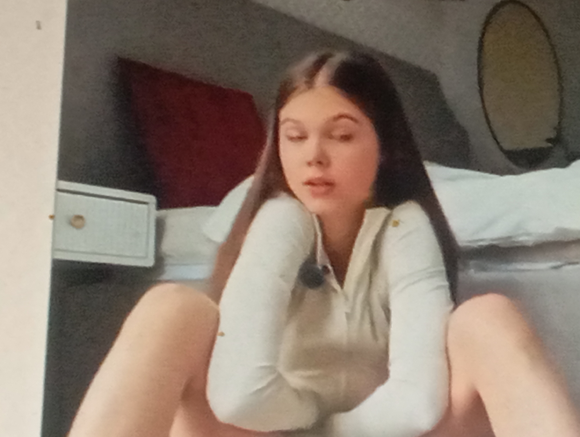 A young person with long, dark hair is sitting on the floor in a room. They are wearing a light-colored, long-sleeved top and their legs are spread. In the background, a bed with white bedding is visible, as is a dark, oval-shaped object mounted on the wall. To the left, there is a white nightstand with a drawer. The scene appears to be an indoor residential setting.