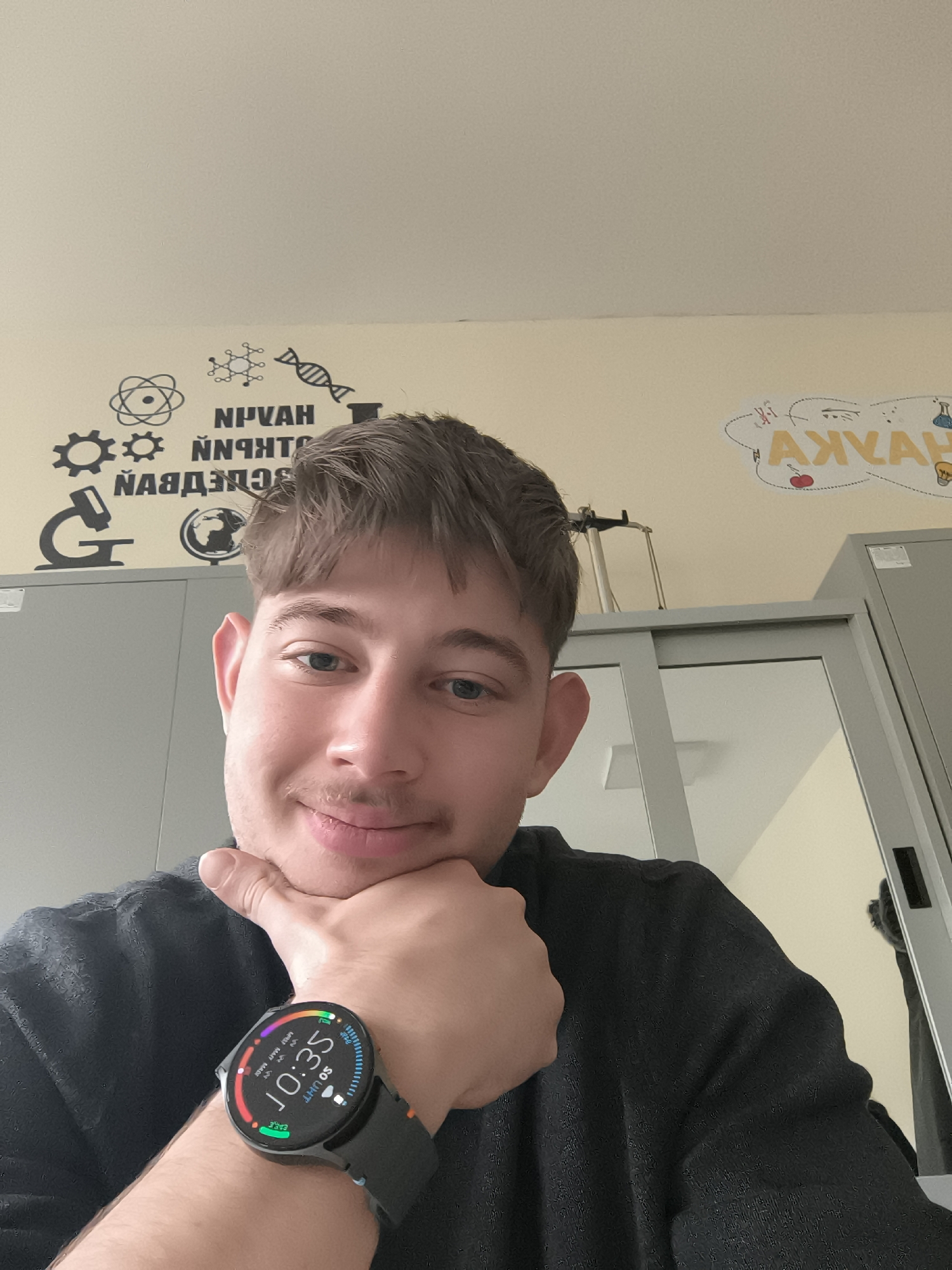 A young man with short brown hair and a slight mustache is looking at the camera, resting his chin on his hand. He is wearing a dark shirt and a smartwatch displaying the time "10:35." Behind him, there are grey cabinets and a light-colored wall with black decals of scientific symbols and lettering in Cyrillic. Additionally, there is a cloud-shaped drawing with Cyrillic letters inside. While the exact location is not discernible solely from this image, the Cyrillic alphabet strongly suggests the city of Sofia, Bulgaria, or another place where the Cyrillic alphabet is used.