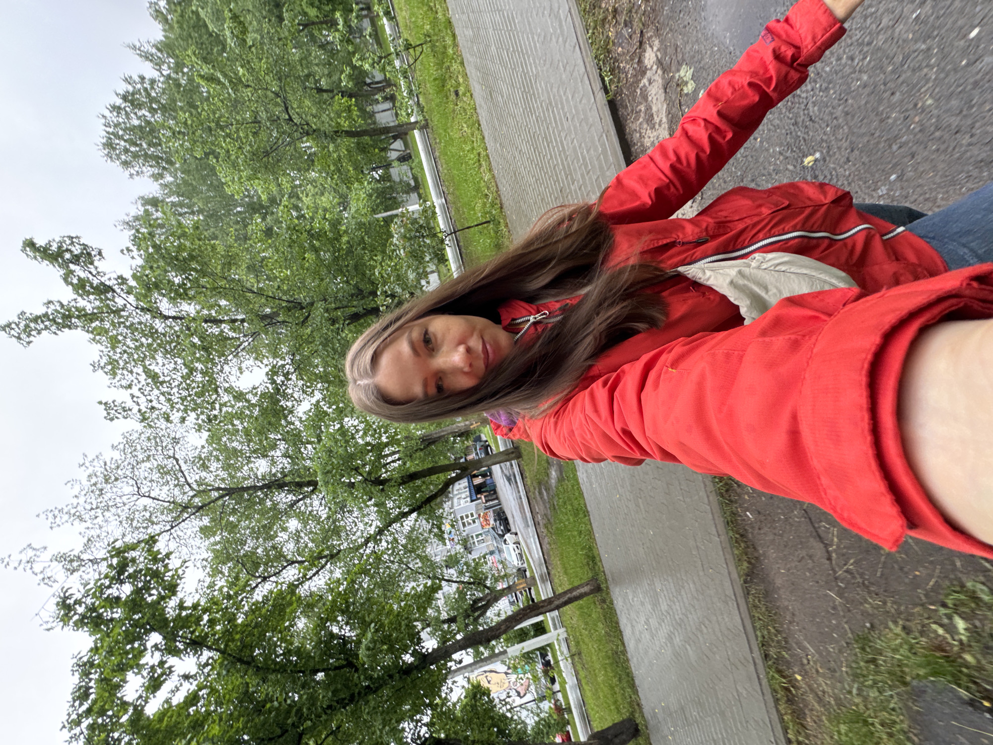 A selfie taken outdoors in Chaikovsky, Russia. A woman wearing a red jacket is in the foreground, with trees and a sidewalk visible behind her. Buildings and cars can be seen in the background across the street. The ground appears wet, possibly from rain.