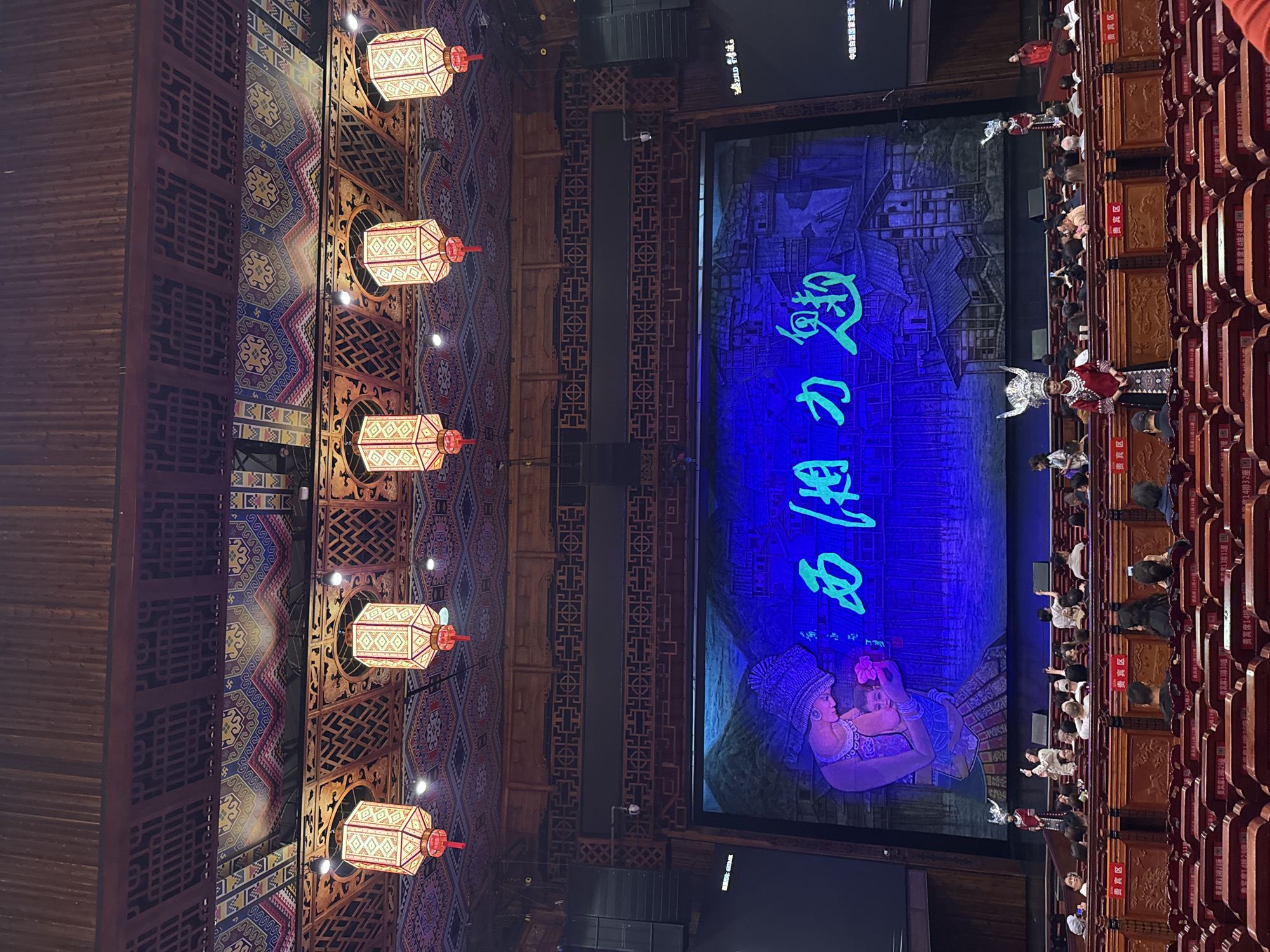 This image captures an indoor cultural performance or show, likely in Wulingyuan, China, given the traditional aesthetic. The scene is dominated by a large, brightly lit stage or screen on the right, displaying a vibrant blue background with green Chinese characters that appear to read "西湘力趣" (Xī Xiāng Lì Qù), potentially the name of the show or a significant phrase. The screen also features stylized imagery, including a depiction of a person in traditional attire at the bottom left, and what seems to be an old village or mountainscape behind the text.
In front of the screen and to its right, an audience is seated in rows of intricately carved wooden seats. Several audience members are visible, some observing the stage, others appearing to interact with performers. On the stage itself, near the audience, there are performers in elaborate traditional costumes. One performer, dressed in red and white with a large, ornate silver headpiece, stands prominently facing the audience. Another performer in similar attire is partially visible further back.
The ceiling of the venue, visible on the left side of the image, is richly decorated with traditional Chinese patterns and wooden lattice work. Hanging from the ceiling are five illuminated square-shaped lanterns, each with intricate designs in red and gold. These lanterns add to the cultural ambiance.
The setting appears to be a formal performance space, likely an evening event, given the controlled lighting that highlights the stage and lanterns while keeping the rest of the hall dim. Text visible on signs attached to the seats and walls indicates "贵宾区" (Guìbīn Qū - VIP area) and specific seat numbers like "贵宾席13排30座" (VIP seat, row 13, seat 30), and "贵宾席14排34座". Some Chinese text is also seen on the upper part of the screen, including "中国白酒国家宝" (China Baijiu National Treasure), possibly a sponsor or theme element. The overall impression is one of a vibrant cultural spectacle.