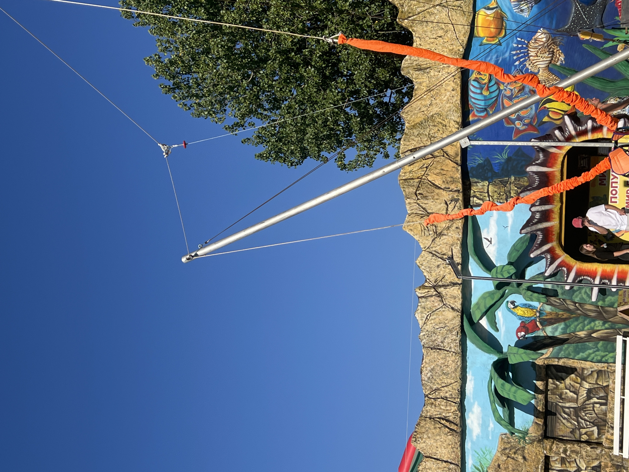 The image depicts an outdoor scene in Golubitskaya, Russia, featuring a bungee trampoline attraction. A tall metal pole, part of the trampoline apparatus, extends diagonally upwards against a clear blue sky. Orange elastic cords hang from the pole, ready for use. In the background, a colorful mural adorns a wall, depicting a jungle scene with palm trees, parrots, and various tropical fish. The mural is set against a rocky textured facade. A few people are visible near the entrance of the attraction, one a woman in a black shirt and another in a white t-shirt and red cap. The overall impression is of a sunny day at a recreational facility.