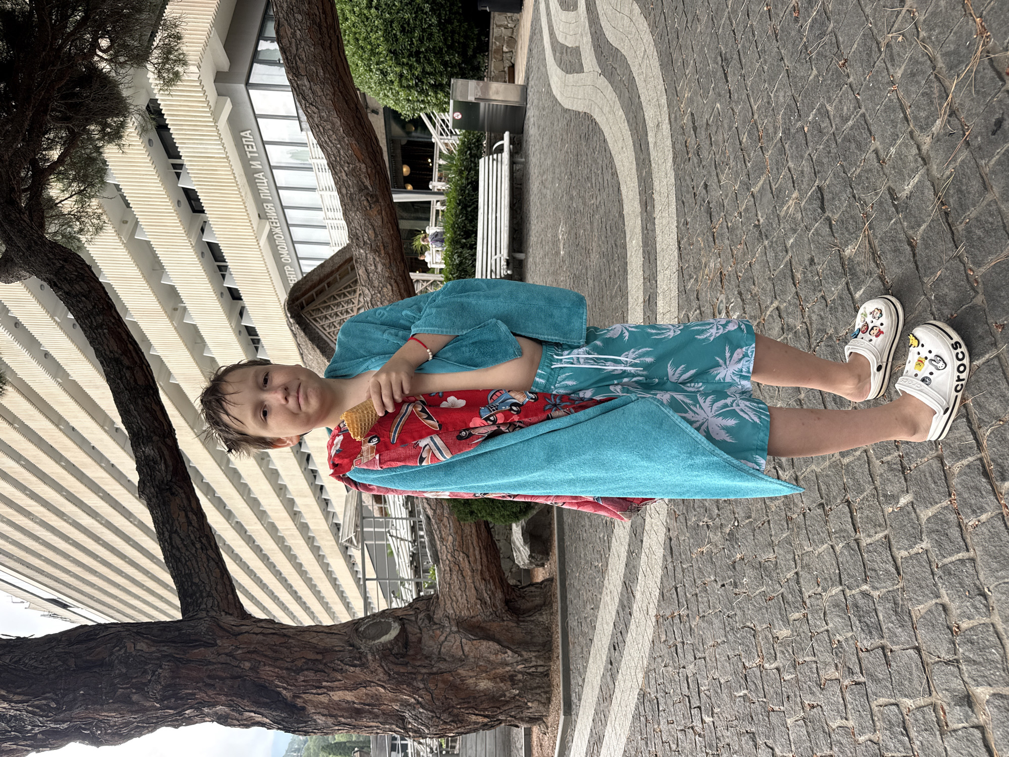 A young boy, wearing patterned swim shorts and white Crocs, stands outside a building in Yalta, Ukraine, with a teal towel draped over his shoulders and a red towel wrapped around his waist. He holds a partially eaten ice cream cone in a waffle cone and has a red bracelet on his left wrist. Behind him, a large, gnarled tree trunk with a branch extending horizontally across the frame, is visible in front of a multi-story building with cream-colored vertical louvers. The ground is paved with gray cobblestones arranged in a curved pattern with white stripes. Several benches and some greenery are also present in the background.