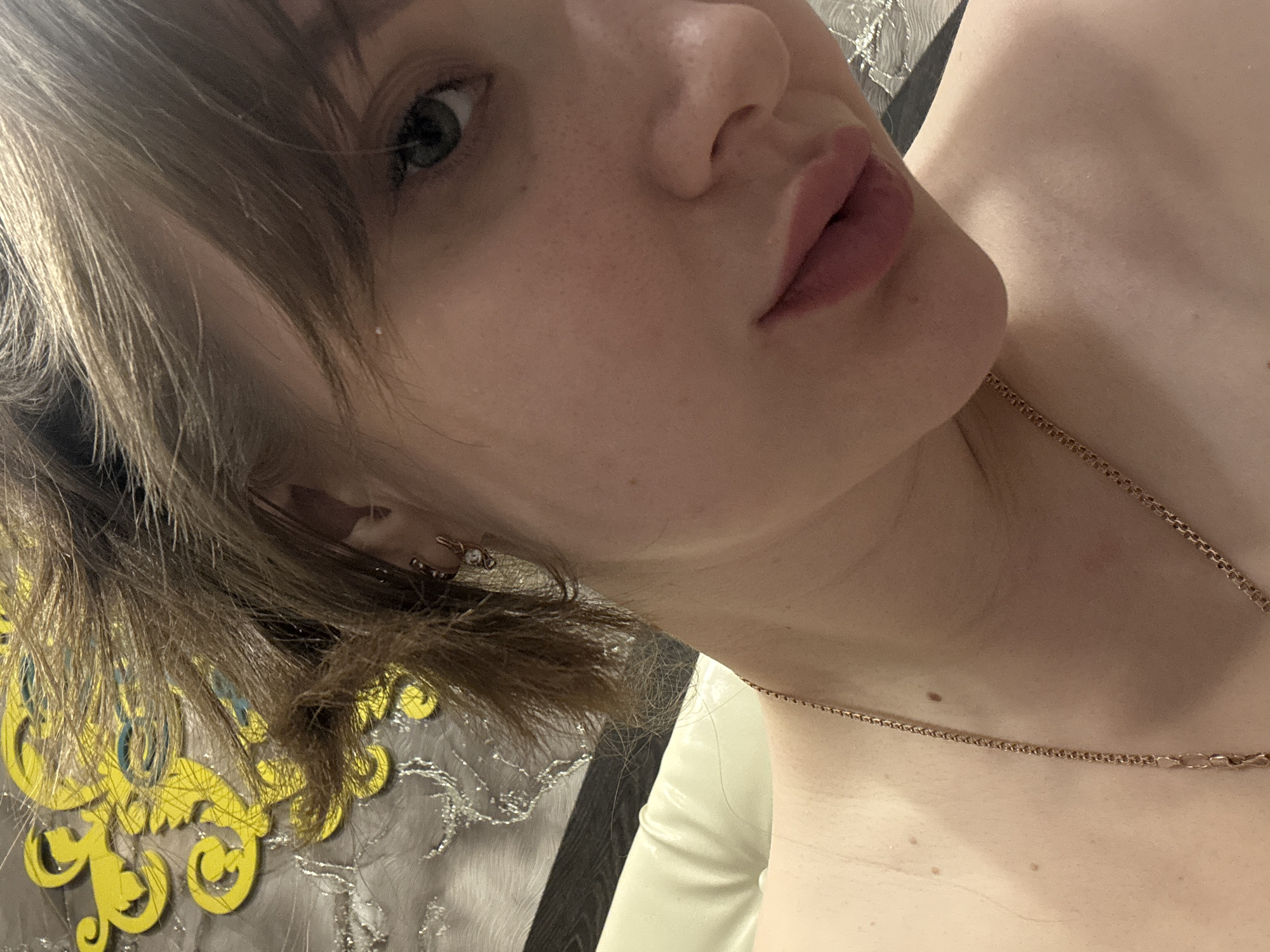 A close-up, angled shot captures the upper portion of a fair-skinned individual. The focus is on their face, showing their left eye with green hues, a slightly upturned nose, and lips with a muted pink color. The person has short, light brown hair, some strands of which frame the face. An earring is visible in the left ear. A delicate gold chain necklace adorns the neck. The background is partially obscured, but elements of a decorative object with yellow and blue flourishes can be seen, along with grey fabric. The location in the city of Chaikovsky, Russia is not discernible from the image.