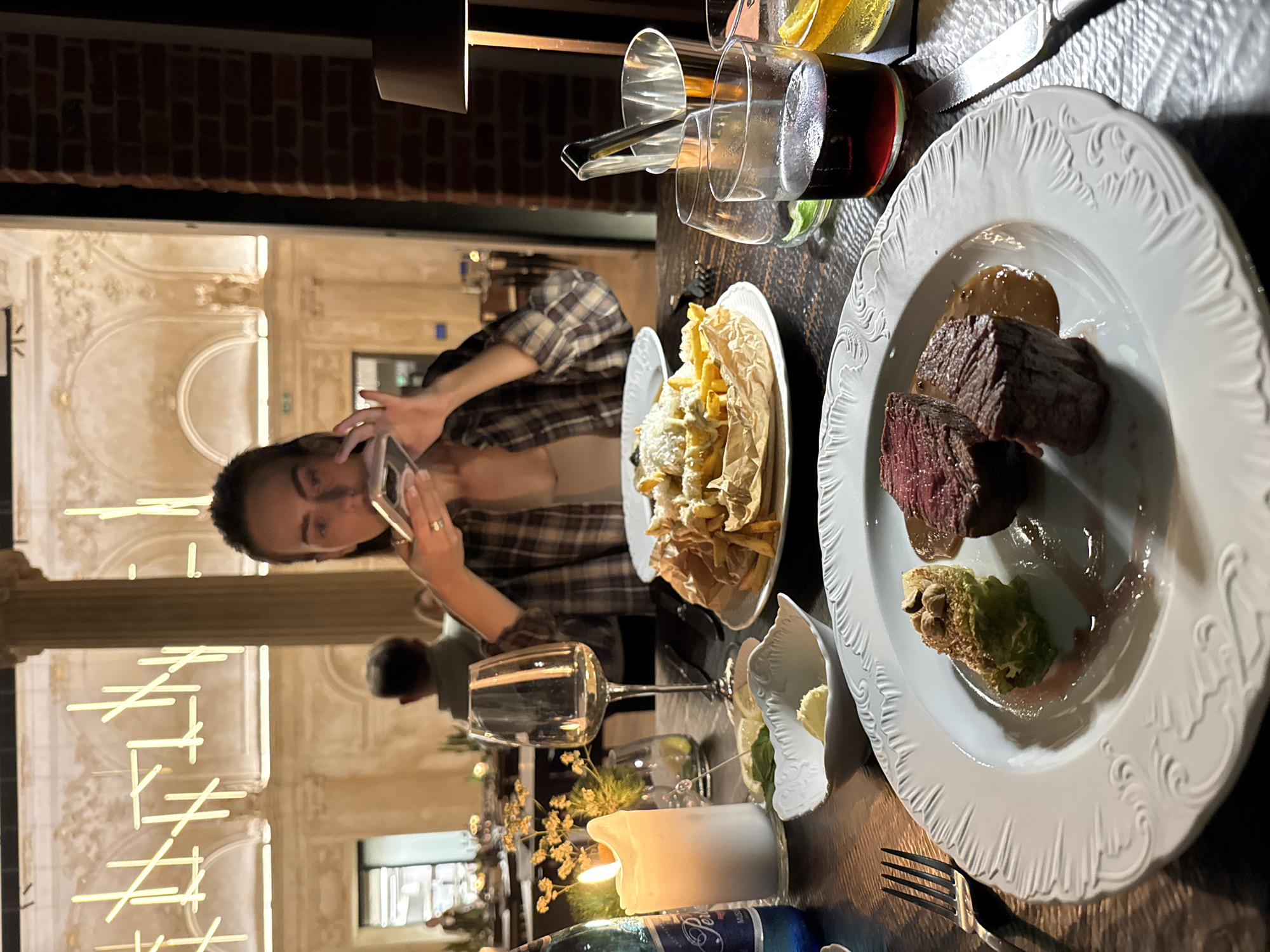 This image captures a nighttime dining scene in an upscale restaurant located in the city of Unknown, Unknown. The foreground features a dark wooden table laden with food and beverages. On the left, a white ceramic plate with an embossed rim holds two medium-rare cuts of meat, accompanied by a green, possibly wrapped, vegetable topped with nuts, and a brown sauce. A silver four-tined fork rests beside the plate. Further left, a blue bottle with "Perlage" visible, a lit white pillar candle, a small white bowl containing a yellow substance (likely butter), and a small dish with mint leaves and a lemon wedge are arranged. A clear, empty stemmed wine glass stands behind these items, next to a small vase with delicate yellow foliage.
In the mid-ground, a person, identified as a female adult with dark hair, is seated wearing a brown plaid shirt over a lighter garment. Her face is partially visible as she holds a silver-colored smartphone up, seemingly capturing an image or video, with a ring visible on her right hand's ring finger. To her right, a second white plate with an embossed rim holds a generous serving of french fries, some topped with a white sauce or cheese, presented on brown parchment paper. Adjacent to this, a short glass tumbler containing a dark red liquid and another with a yellow liquid and an orange slice are visible, alongside a silver knife and a small silver ice bucket with tongs. A table lamp with a dark shade casts light onto the right side of the table.
The background reveals an expansive, elegantly lit interior space characterized by high ceilings, a large stone column, and multiple arched doorways or niches with ornate detailing. Modern, abstract light fixtures, composed of glowing white/yellow horizontal and diagonal bars, adorn the walls. A male figure wearing a green/grey hooded sweatshirt is seated in the background, mostly obscured. The rightmost edge of the image shows a section of a dark red brick wall. The overall ambiance is dimly lit, suggesting an intimate or sophisticated dining experience.