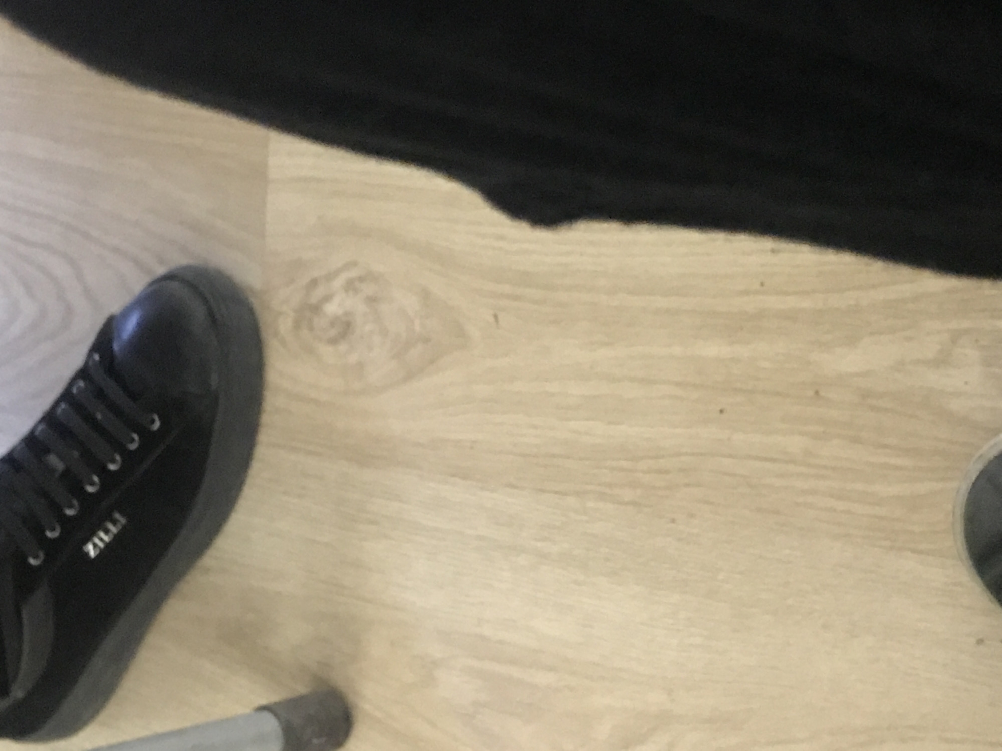 The image shows a floor covered in light wood-look planks, partially obscured by black clothing. A black "ZILLI" branded sneaker is visible in the lower left corner. A section of gray metal pipe is adjacent to the sneaker. Another shoe, partially visible in the lower right, is gray. The location is not apparent.