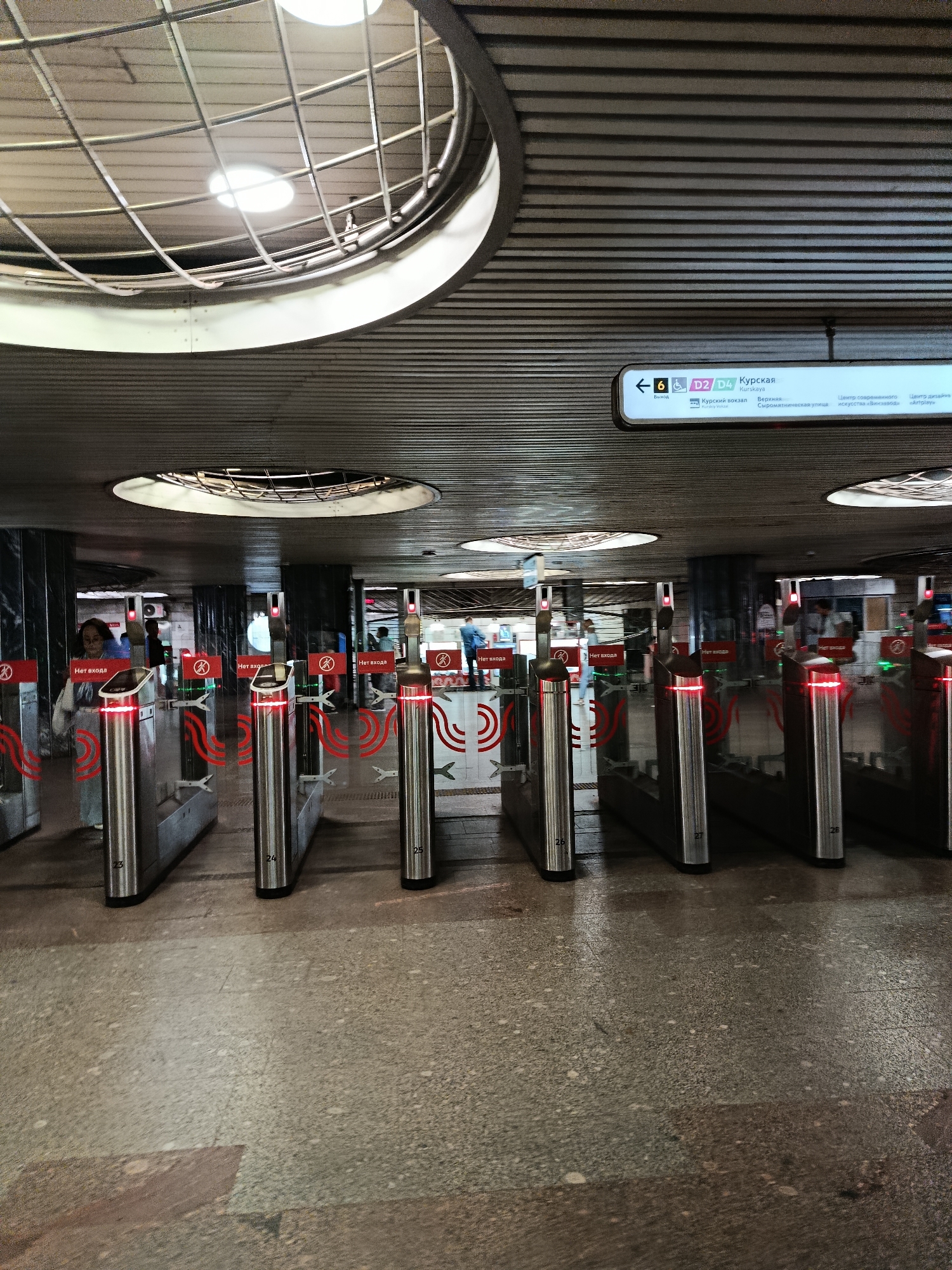 The image depicts the interior of a Moscow Metro station, likely Kurskaya, given the signage. The scene is characterized by a modern, utilitarian aesthetic with a focus on functionality.
The foreground is dominated by rows of silver turnstiles, each equipped with red indicator lights and some displaying accession numbers like "23", "24", "25", "26", "27", and "28". Above the turnstiles, red signs with a circle and a diagonal line, likely indicating "No Entry", are visible. In the background, a few people are seen passing through the turnstiles, suggesting the station is operational.
The ceiling features a distinctive ribbed metal design with circular light fixtures. A prominent sign above the turnstiles provides directional information, indicating lines "6", "D2", and "D4" and the destination "Kurskaya". It also lists destinations such as "Kursky vokzal" (Kursky train station) and "Syrtyanitheskaya ulitsa" (Syrtyanitheskaya Street), along with more cultural destinations like "Center of contemporary art 'Vinzavod'" and "Artplay".
The floor appears to be paved with speckled granite tiles. The overall impression is one of a busy, efficient public transportation hub. The lighting is bright and functional, illuminating the passageways and equipment. The architecture suggests a contemporary design within the older infrastructure of the Moscow Metro system.