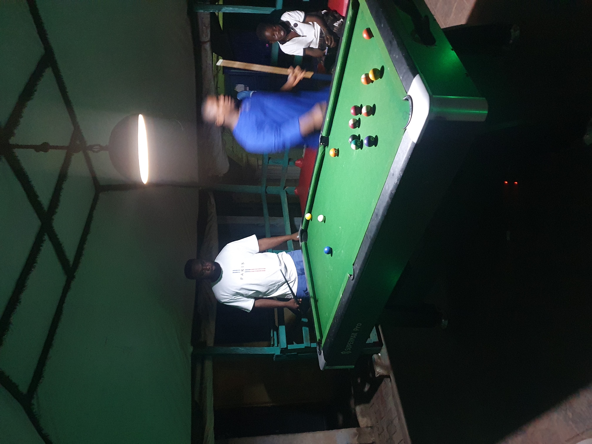 In Lokoja, Nigeria, a dimly lit, open-air pavilion features a green billiard table, illuminated by a hanging lamp and under-glow lighting. Three individuals are present: a man in a white "Paris" t-shirt and blue shorts stands on the left, holding a billiard cue; a second man, wearing a blue shirt, is blurred in motion on the right, also holding a cue; and a young person in a white collared shirt sits on a red bench in the background. Several billiard balls are scattered across the table. The pavilion's ceiling is covered with a light-colored fabric, crisscrossed by dark beams, and the surrounding structure appears to be made of wood and metal. The ground is paved with dark bricks.