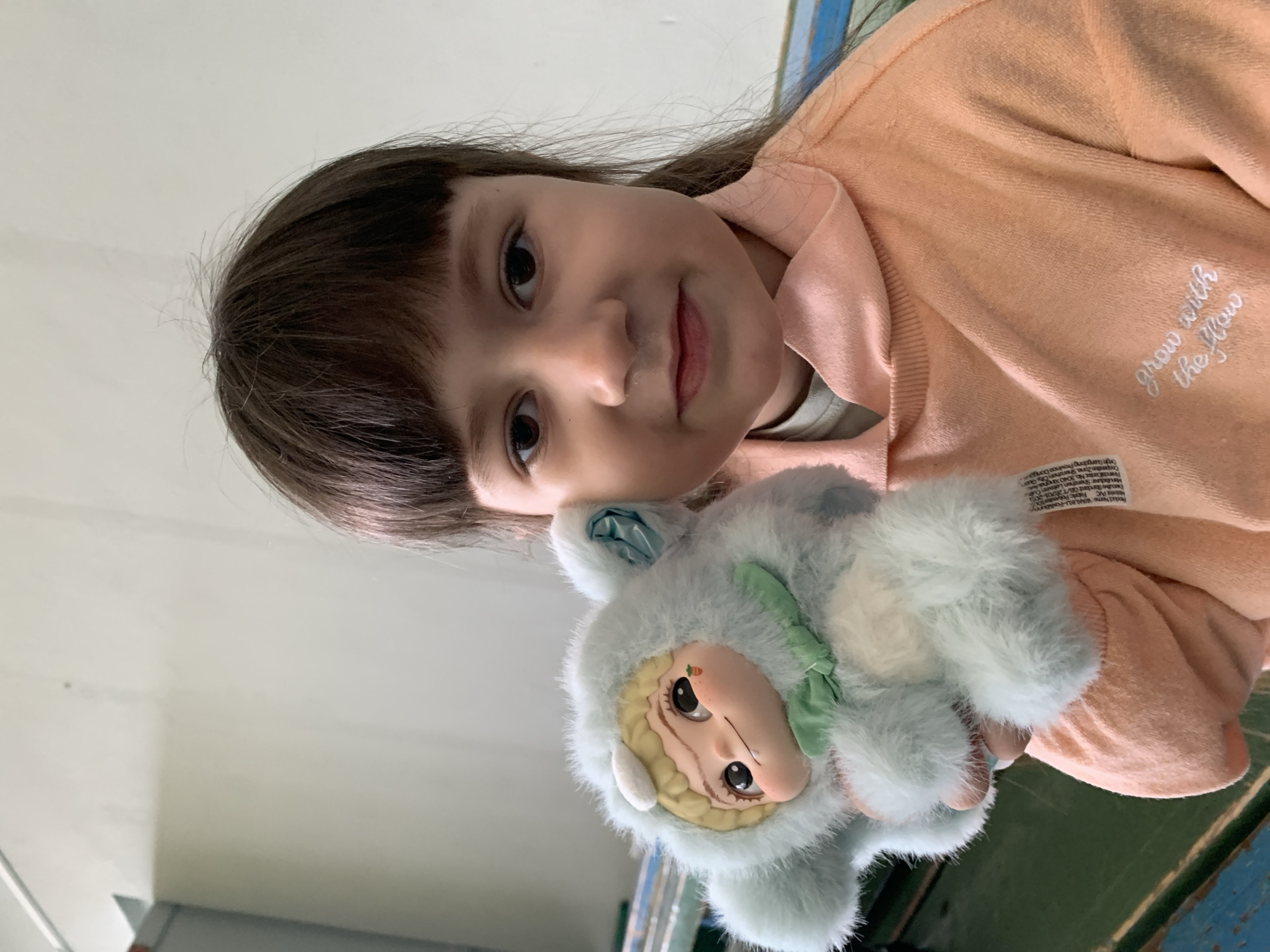 A young girl with brown hair and bangs is holding a plush toy. She is wearing a light orange sweatshirt with the phrase "grow with the flow" embroidered on it. The plush toy is light blue and white with a tan face and a green bow. The background consists of a white wall and a blue and green surface. The OCR text references "WAALIKLA" and a location in Dongguan City, Guangdong Province. The image does not contain information about the city of Chaikovsky, Russia.