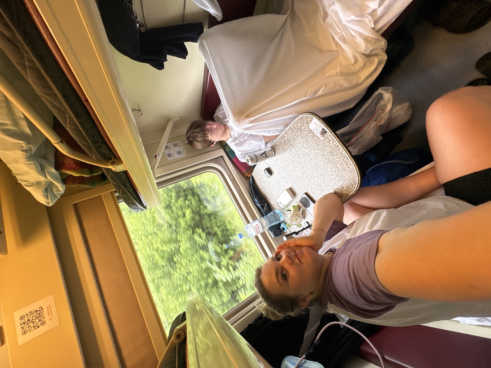 The image depicts the interior of a train compartment, with the window revealing a blur of green trees indicating movement. In the foreground, a woman with blonde hair pulled back is looking towards the camera, resting her chin on her hand. She is wearing a purple and white t-shirt. To her right, a table is visible with a water bottle, a small fan, a smartphone, and a power bank. In the background, a young boy with short brown hair is seated, wearing a white t-shirt and appearing to be looking down. He is partially obscured by a white sheet. The compartment features bunk beds with bedding, and clothing is hung on a rack. A QR code is affixed to the wall above the window. The overall lighting suggests it is daytime. The location is identified as the city of Баграш-Бигра, Russia.