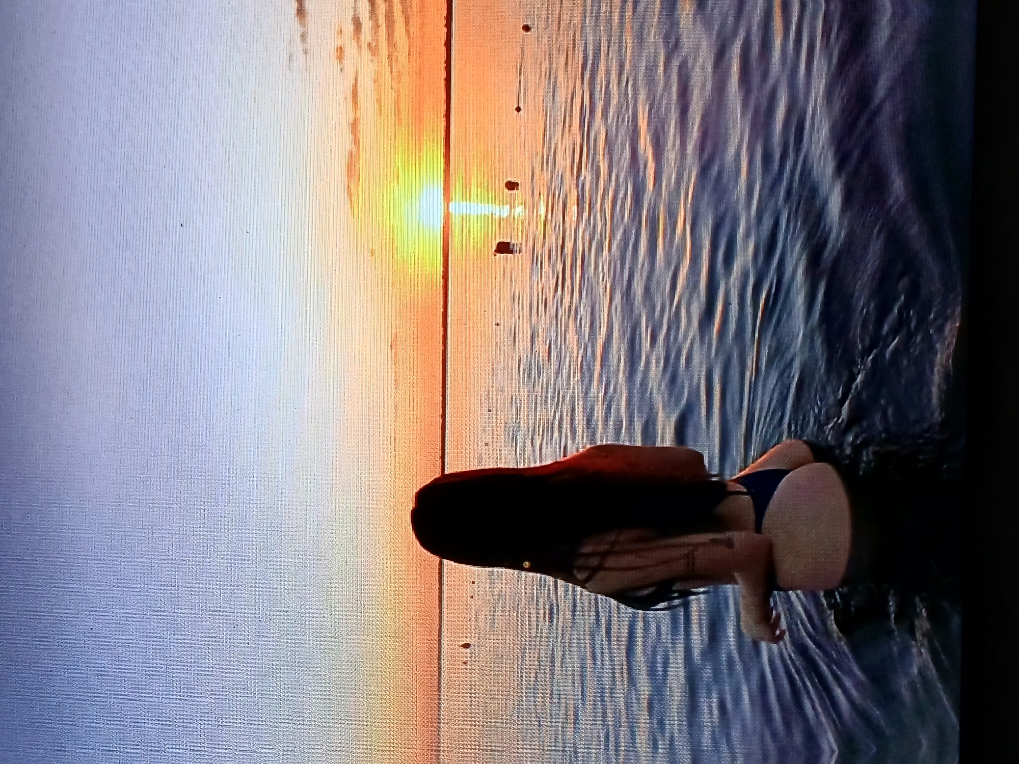 A woman is shown from the back, partially submerged in water near the shore at sunset. The water appears to be shallow, with gentle ripples and the reflection of the setting sun. The sky is a gradient of colors from light blue to orange near the horizon. It is likely in the vicinity of Juan Griego, Venezuela.