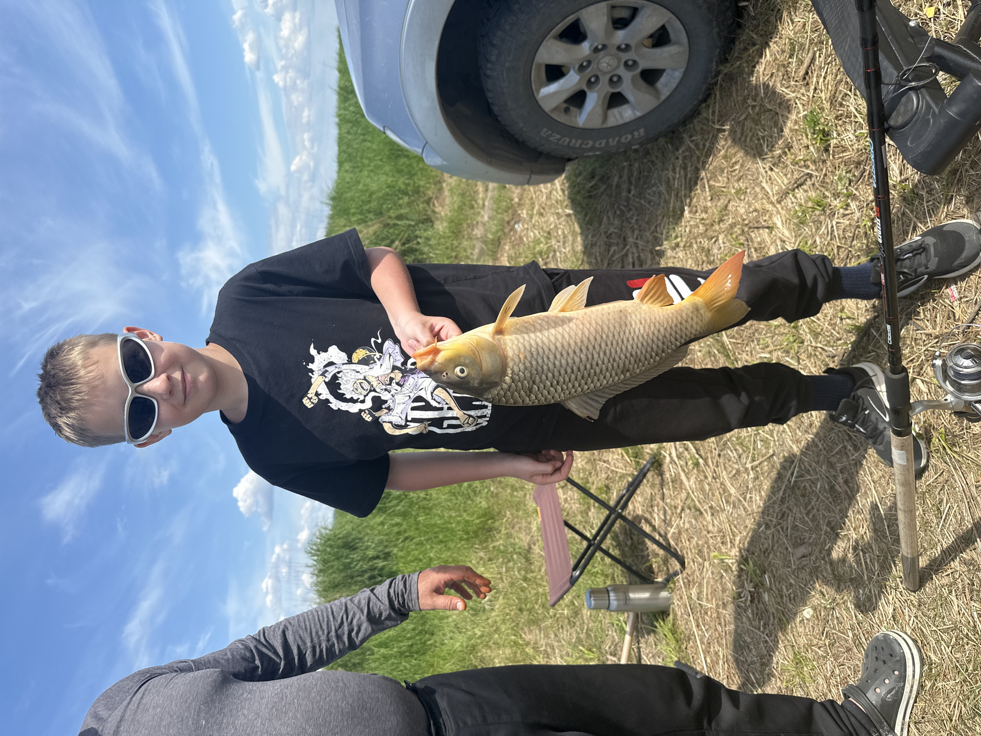 This media file depicts a young boy, likely a teenager, holding a freshly caught fish. He is wearing a black t-shirt with a graphic print, black pants, and white-framed sunglasses. His short, light brown hair is visible. The fish, a large carp with golden scales, is held horizontally in front of him.

The boy is outdoors, in a grassy area with dry hay and some green vegetation. A car, with a tire visible, is parked in the background. A fishing rod and reel are also present, resting on the ground next to the boy. Another person, only their arm and upper body are visible, is in the foreground, wearing a gray long-sleeved shirt.

The scene appears to be a fishing trip, likely on a sunny day given the bright lighting and blue sky with scattered clouds. The overall mood seems positive, with the boy proudly displaying his catch. The text "ROADCRUZA" is visible on the car tire. The location is noted as Im. Tel'mana, Russia.