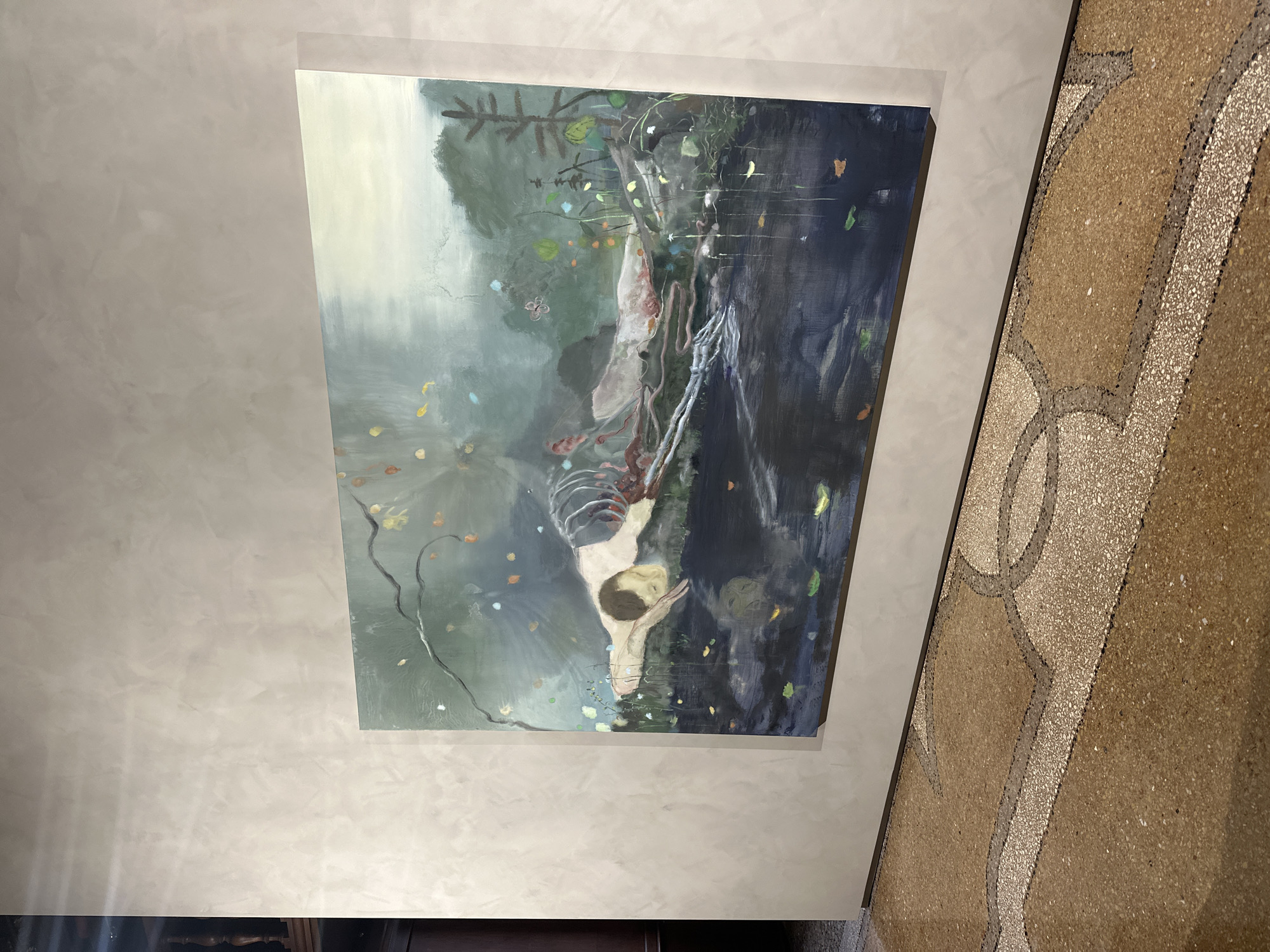 The image shows a painting hanging on a light beige wall. The painting depicts a figure in a watery landscape, with skeletal elements and dark, murky water. The wall has a textured finish. To the right of the painting is a wall or flooring with a patterned terrazzo design in beige and brown tones. The location is not explicitly stated to be Venice, Italy in the image and that information can not be provided.