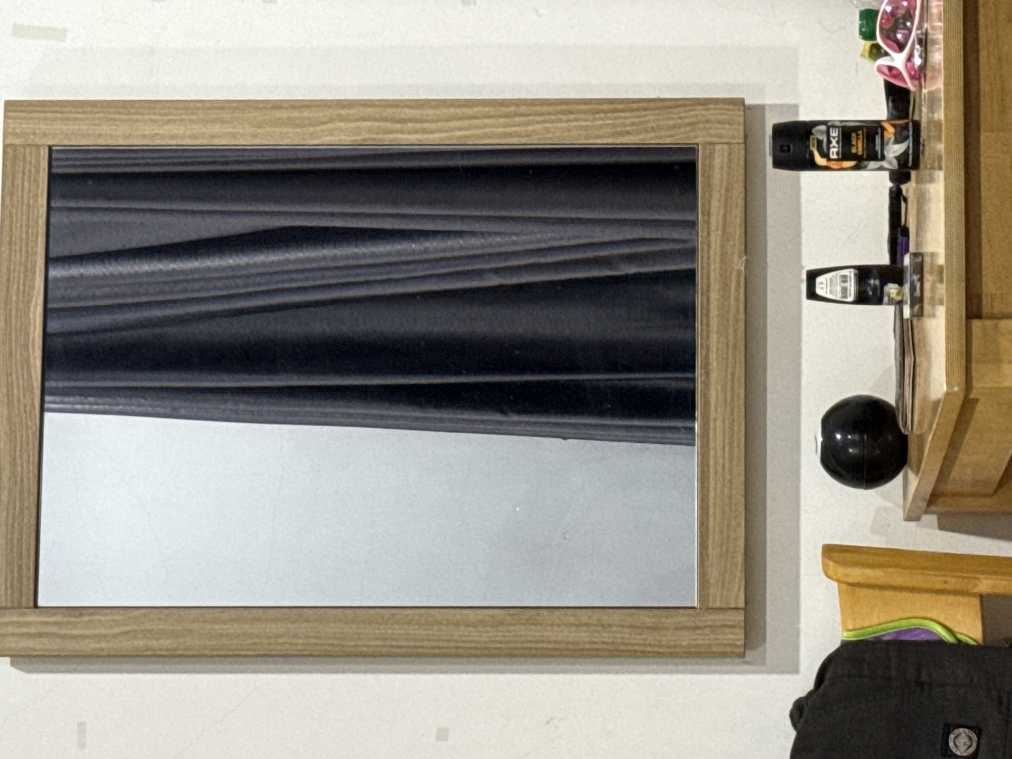 An indoor, eye-level shot captures a rectangular mirror with a light wooden frame affixed to a white wall. The mirror reflects a dark, horizontally undulating surface in its upper half, transitioning to a plain light gray area in the lower half.
To the right of the mirror, a light-colored wooden shelf unit is mounted on the wall. On one of its shelves, two spray cans, likely deodorants, are visible. The upper spray can is black with clear text "AXE BLACK VANILLA" printed on it. The lower can is also dark but its white label is indistinct. Above these cans, on an upper shelf, a pair of pink-rimmed sunglasses and other small items are partially visible.
A glossy black spherical object is attached to the wall near the bottom of the shelf unit. Below this, a small wooden block is seen, and beneath that, a dark gray or black piece of clothing, possibly shorts or pants, with an unreadable circular logo or patch. The white wall displays minor textural variations and marks. No people or active events are depicted. The scene suggests a personal interior space like a bedroom or bathroom. No specific visual cues indicate the location as Đà Nẵng, Vietnam.