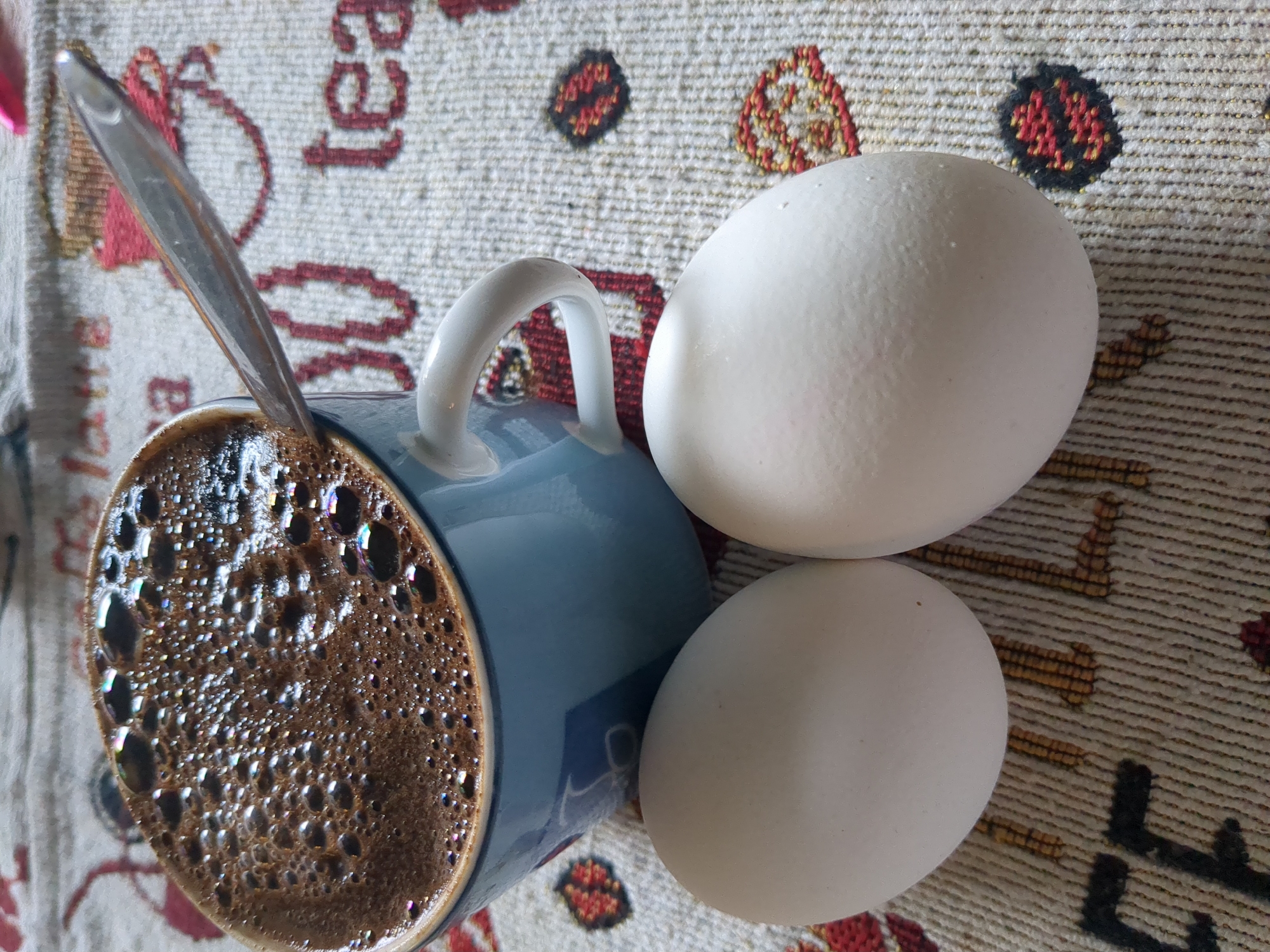 A close-up, high-angle shot shows a light blue ceramic mug filled with dark brown coffee, topped with a thick layer of foam. A silver spoon rests on the rim of the mug, its handle extending out of the frame. Two white eggs lie in front of the mug on a tan, textured placemat. The placemat has a repeating pattern of red and black circular designs. The image is well-lit, with the light reflecting off the coffee and spoon. The focus is sharp on the coffee, spoon, and eggs. The background is slightly blurred, showing more of the patterned placemat. There are no people or animals visible in the image. The scene appears to be set on a table indoors. No specific location in Nordhausen, Germany, can be identified from this image.