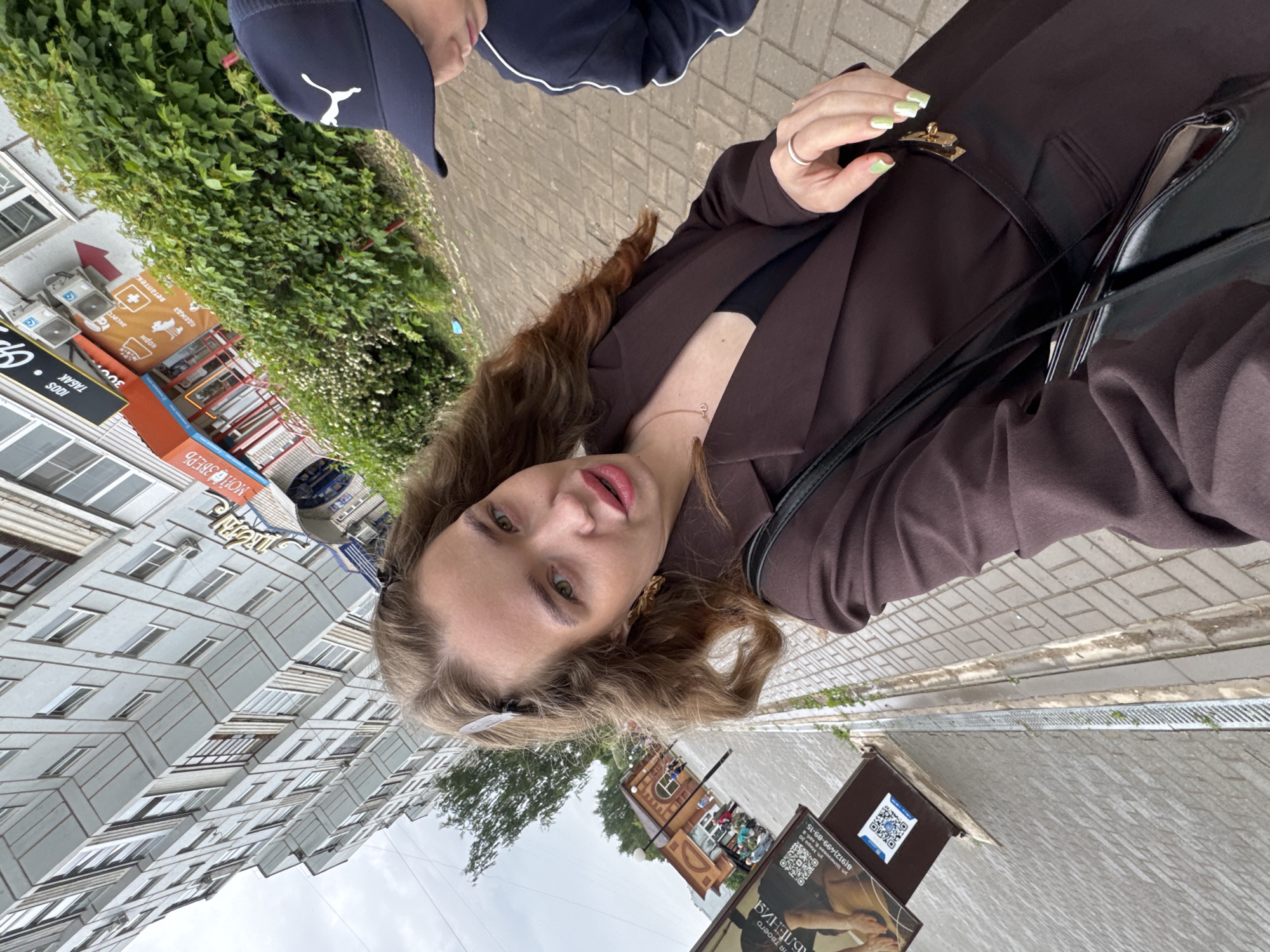An outdoor, slightly tilted selfie captures a young woman and a street scene in Chaikovsky, Russia, under an overcast sky during daytime. The primary subject is a woman with long, wavy light brown hair, green eyes, and pink lips, looking directly at the camera. She wears a dark brown blazer over a black top, a delicate gold necklace, a silver ring, and has light green and white painted nails. She carries a black shoulder bag with a gold clasp. Partially visible behind her to the left is another person wearing a dark blue cap with a white Puma logo.
The background features a grey interlocking brick pavement and a curb with a storm drain. To the left, a tall, grey multi-story apartment building with numerous windows and balconies rises. Storefronts with various signs, including "ТАБАК" (Tobacco), "аксесс" (accessories), and "МОЙ ЗВЕРЬ" (My Beast/Pet) alongside "корм" (feed/food), "одежда" (clothing), and "Ветаптека" (Veterinary pharmacy), are visible on the ground level of a building. A red arrow sign and air conditioning units are also present. Lush green bushes line the street. Further down the street, an older brown brick building with people gathered outside can be seen. In the foreground, a brown advertising stand displays QR codes and text mentioning "ул. Шлюзова" (Shlyuzova Street) and "ул. Мира" (Mira Street), along with a phone number "8(912)499-89-15" and "АБЛЕНИЯ".