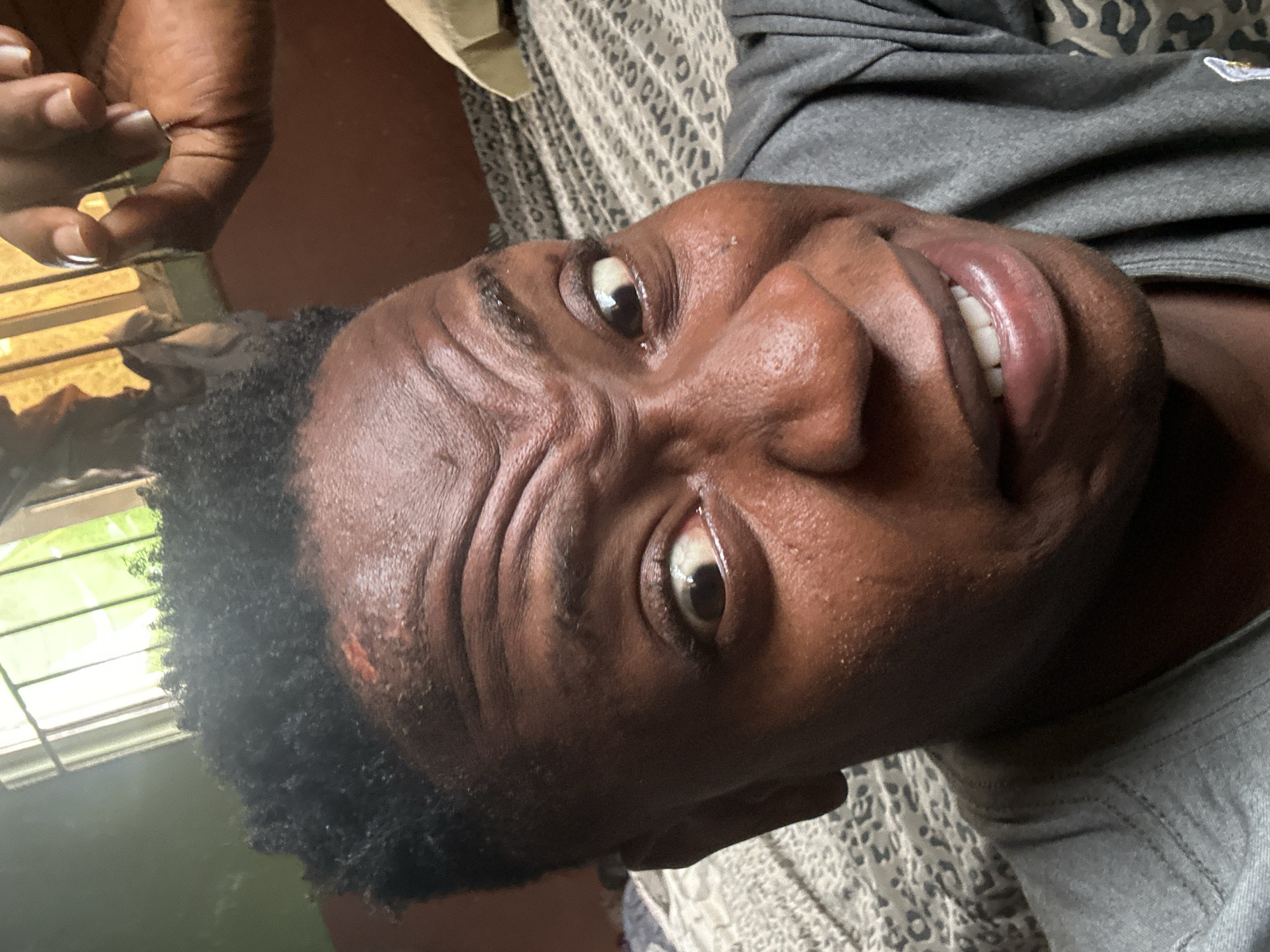 A close-up, selfie-style photograph captures a young Black male with dark skin and short, dark, curly hair. He is wearing a grey t-shirt. His facial expression is one of surprise or alarm, with wide, staring eyes and furrowed eyebrows. His mouth is slightly open, revealing his teeth. The background shows a patterned bedspread and a window with visible green foliage and what appears to be metal bars or a grille. A hand with manicured nails is visible in the upper right corner of the frame, reaching towards the subject. The lighting suggests an indoor setting, possibly daytime. The image appears to have been taken in Ikorodu, Nigeria, indicating a local context within a specific urban environment.