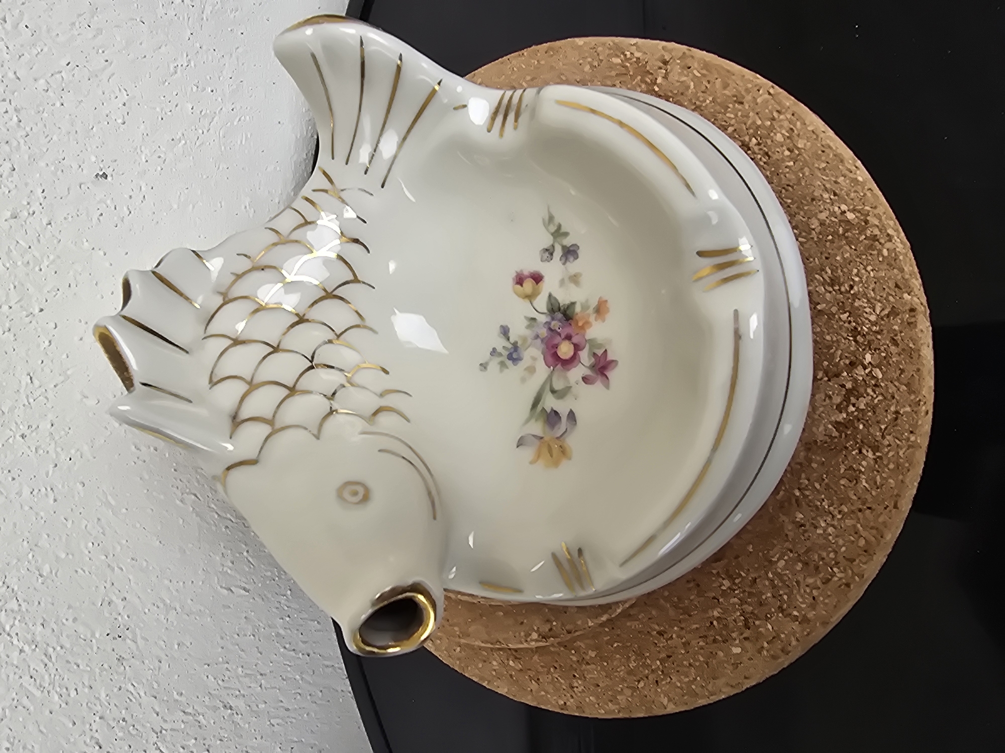 A ceramic fish-shaped ashtray is resting on a circular cork coaster. The ashtray is white with gold accents and features a floral design inside the bowl. The head of the fish has a gold-rimmed opening. The cork coaster sits on a dark, possibly black, surface. A textured white wall is visible to the left of the ashtray. The location of the scene is Unknown, Unknown.