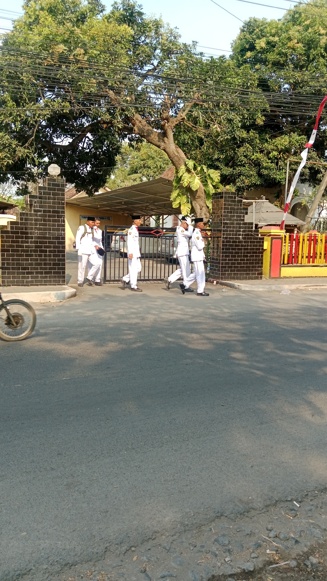 A group of five young men in white uniforms and black caps are walking in a line towards the right side of the frame. They appear to be students, possibly on their way to or from an event or school. The men are walking on a paved road in front of a black brick wall with a metal gate. Behind the gate, there is a building with a metal roof. The environment appears to be a residential or school area in Patunjungan, Indonesia, with trees and a clear sky. A bicycle wheel is visible in the lower left corner of the image. The overall scene suggests a typical day in a town in Indonesia.