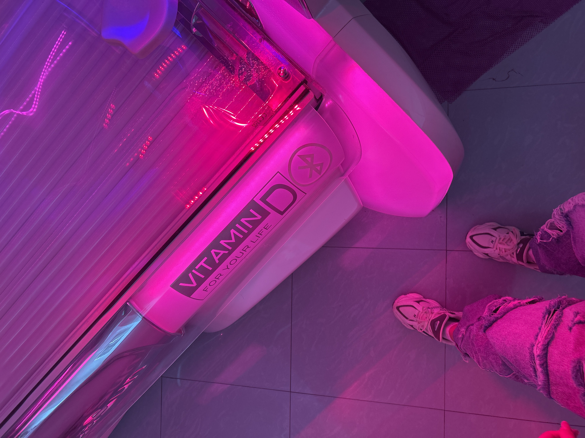 A top-down shot depicts a tanning bed under bright pink and blue lighting. The tanning bed features the words "VITAMIN D" in a prominent font on one of its sides. The lower portion of a person stands to the right of the tanning bed, wearing sneakers and ripped jeans. The setting appears to be indoors, possibly a tanning salon, and the floor consists of tiled squares.
Lovech, Bulgaria is not mentioned in the image.