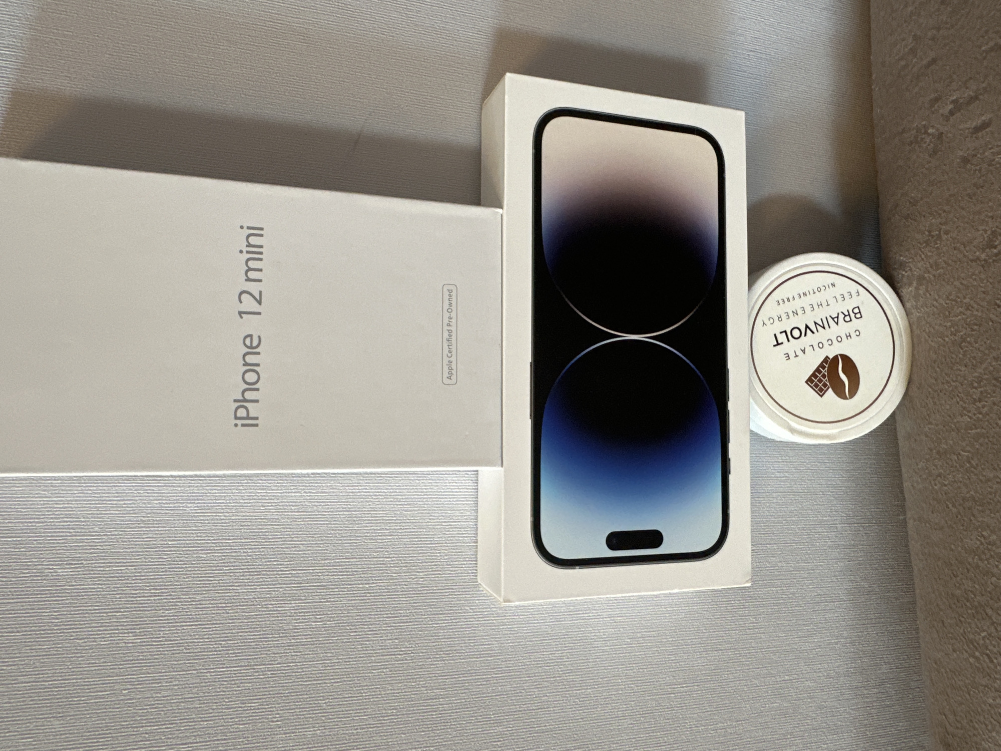 An iPhone 12 mini box is stacked on top of a larger box displaying a smartphone screen with a blue and purple gradient. Below the smartphone box is a small, round container labeled "BRAINVOLT CHOCOLATE FEEL THE ENERGY NICOTINE FREE". The items are positioned against a textured, light-colored wall and resting on a fabric surface. The lighting suggests an indoor setting, possibly a room or display area. No specific geographical indicators for Birobidzhan, Russia, are visible.