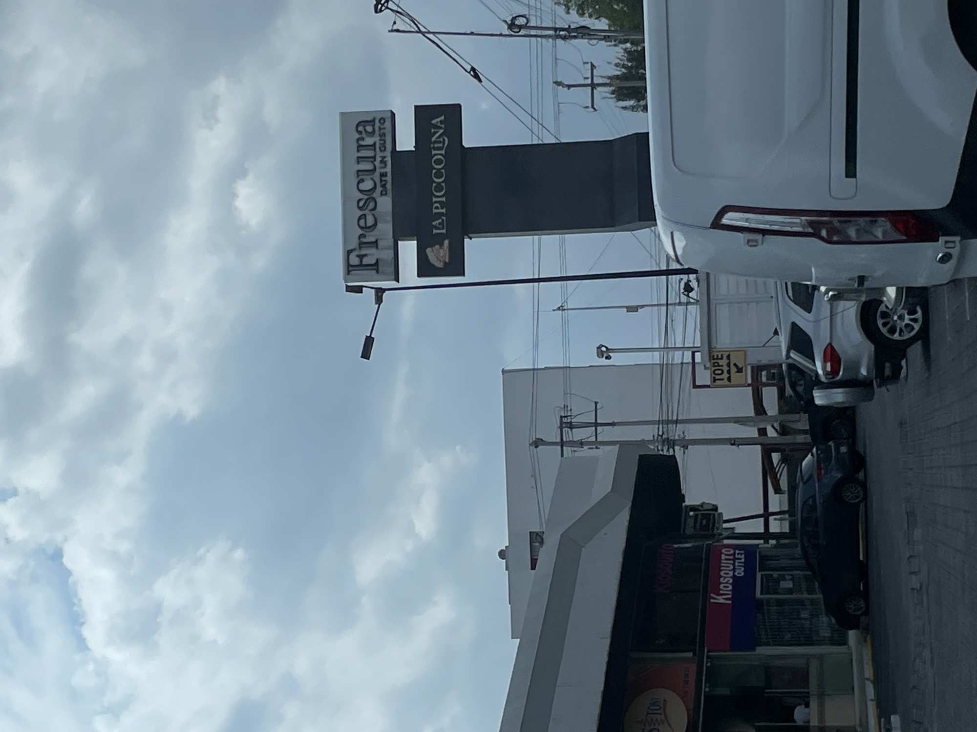 The image shows a street scene in Puebla, Mexico. A sign for "Frescura Date Un Gusto" and "La Piccolina" is visible. There's a white van parked on the street, and other cars are parked or driving nearby. A building with "Kiosquito OUTLET" signage is also present. The sky is overcast. Overhead power lines and utility poles are part of the street environment.