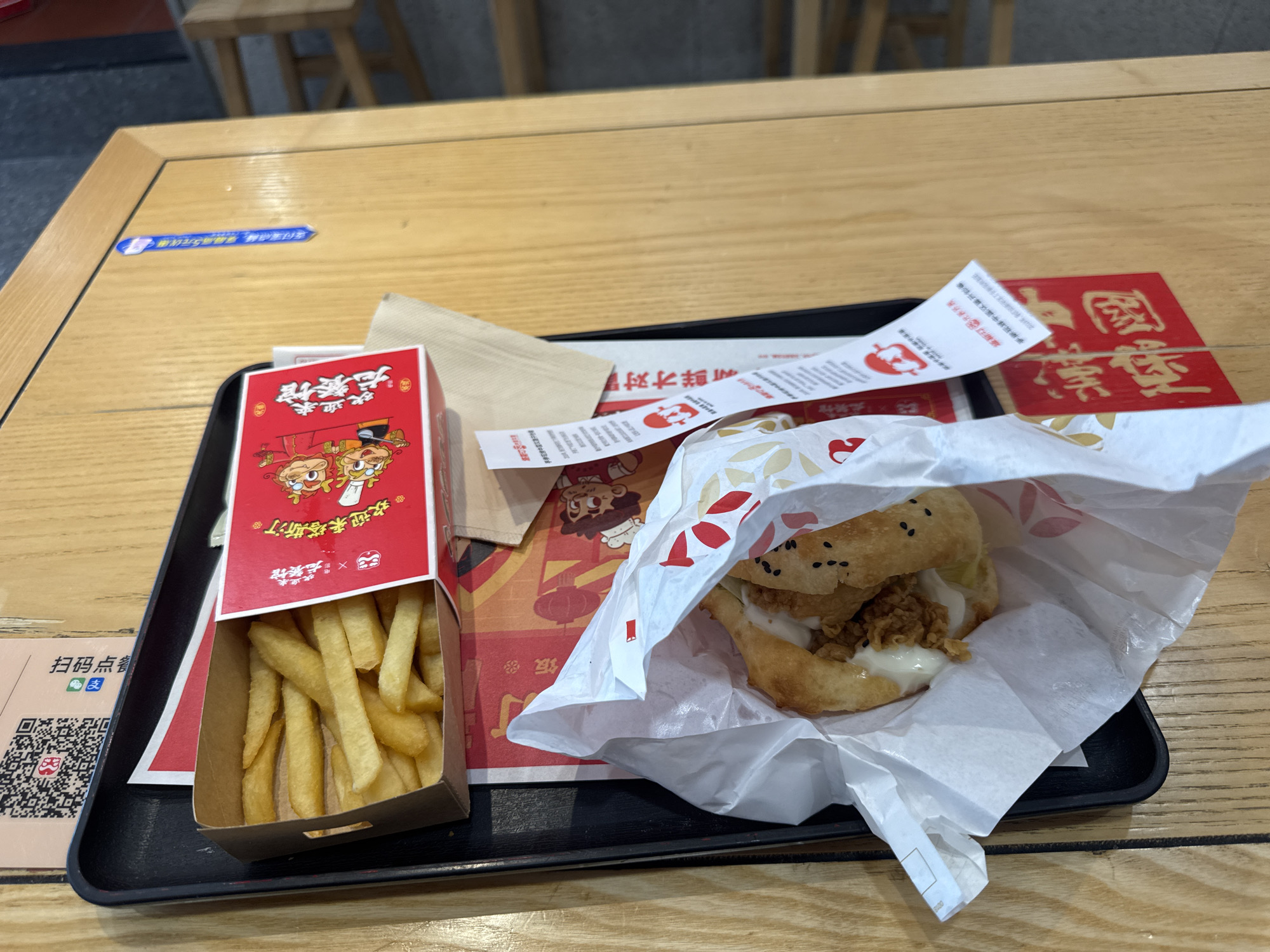 A close-up, eye-level, outdoor shot shows a fast-food meal on a wooden table at a restaurant in Dadong District, China. The meal consists of a burger, french fries, and what appears to be a beverage cup partially visible in the background. The food is served on a black tray. The burger is wrapped in white paper, with part of it peeking out. The french fries are in a red and white cardboard box with cartoon characters on it. There are also some paper wrappers and napkins on the tray. In the background, a QR code and some Chinese text are visible on a light brown surface. The overall scene suggests a casual dining experience.