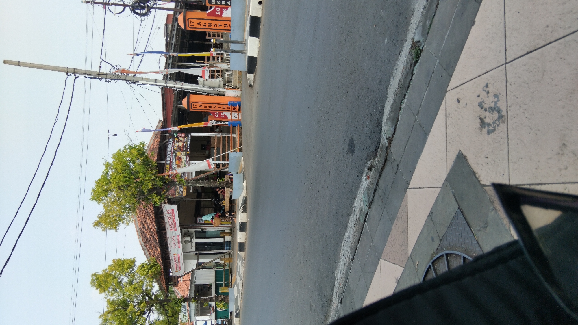 The image shows a street scene in Batang, Indonesia. On the right side of the image, there is a building with an orange awning and flags. The building appears to be a shop or restaurant. There are several people visible, some sitting at tables outside the building and one person walking past. The street is paved with asphalt, and there is a sidewalk on the left side of the image. The sky is clear and the sun is shining. The location appears to be a commercial area, with several businesses visible. The flags suggest a celebration or event is taking place.