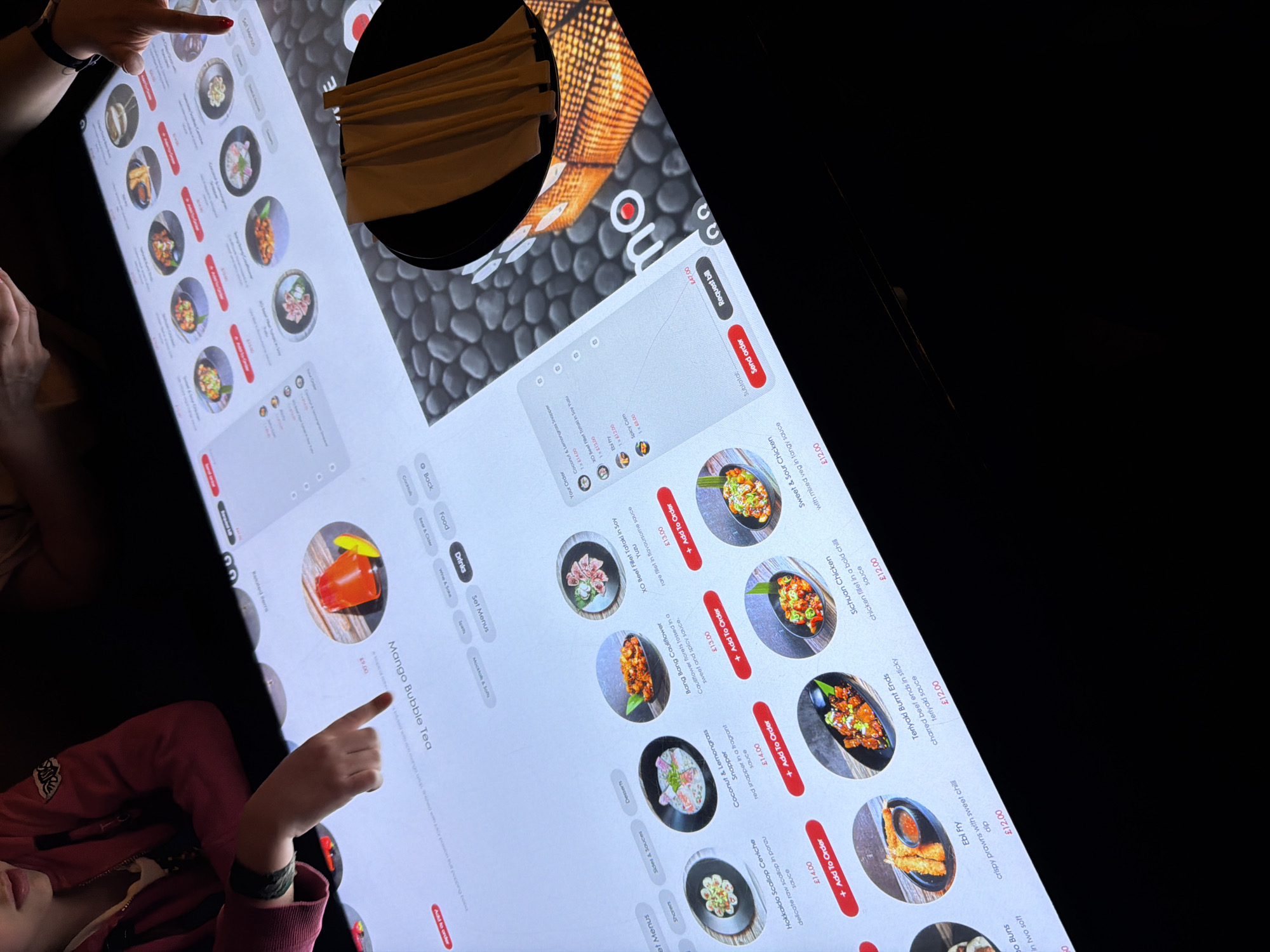 The image captures a brightly lit interactive touch-screen menu at a restaurant, likely located in the city of London, United Kingdom. The angled perspective shows a section of the menu, listing various Asian-inspired dishes such as "Sweet & Sour Chicken," "Ebi Fry," and "Mango Bubble Tea" along with their prices in pounds sterling (£). An adult's finger is seen pointing at the top of the menu, while a child's finger is pointing toward the "Mango Bubble Tea." A separate section displays the order summary, along with buttons to request the bill or send the order. The restaurant's logo "mo" is visible in the upper right. A container filled with chopsticks can be seen next to the menu.